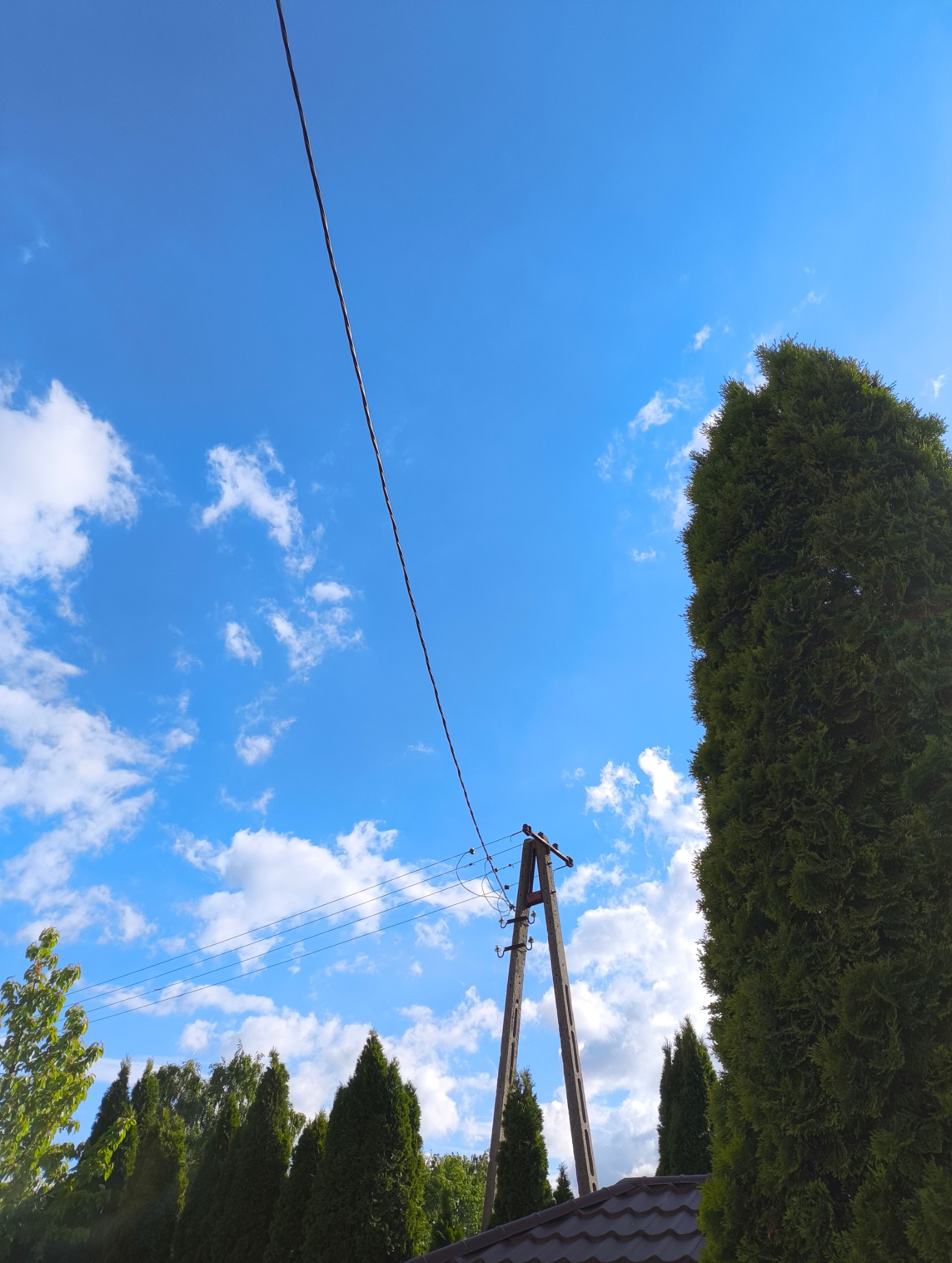 Słup energetyczny z liniami wysokiego napięcia na tle błękitnego nieba z chmurami i zielonych drzew. Widok z dołu, na pierwszym planie fragment dachu.