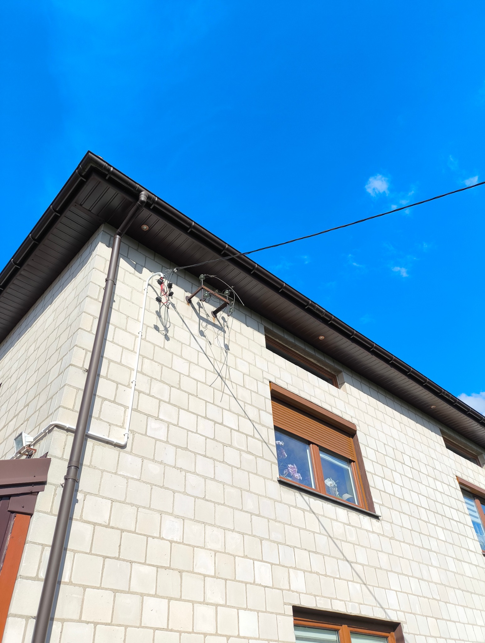 Narożnik domu z białej cegły z widoczną instalacją elektryczną na ścianie i brązowym orynnowaniem na tle błękitnego nieba.