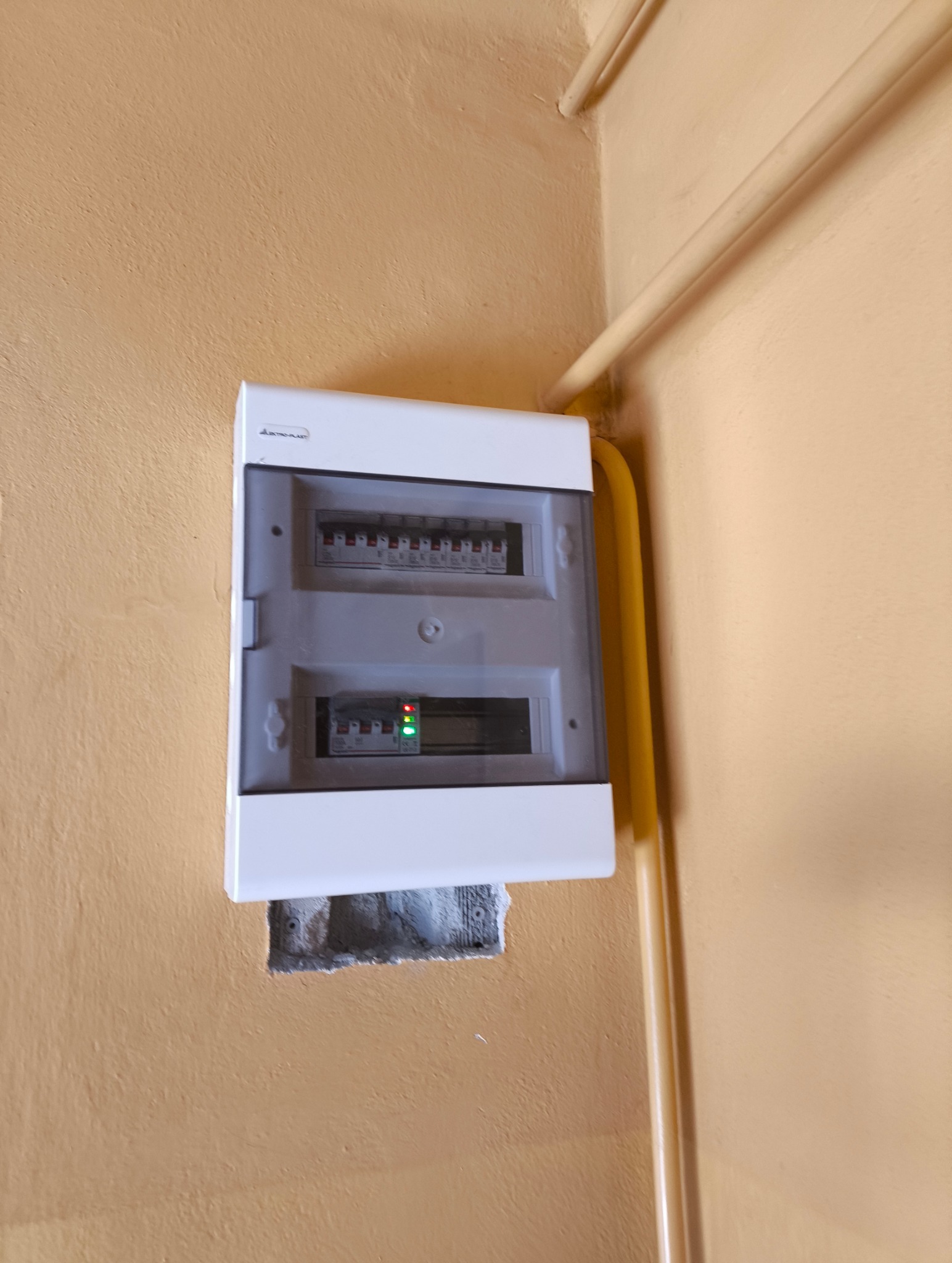 Zamknięta skrzynka elektryczna z widocznymi bezpiecznikami i diodami, zamontowana na pomarańczowej ścianie w rogu pomieszczenia, obok żółtej rury.