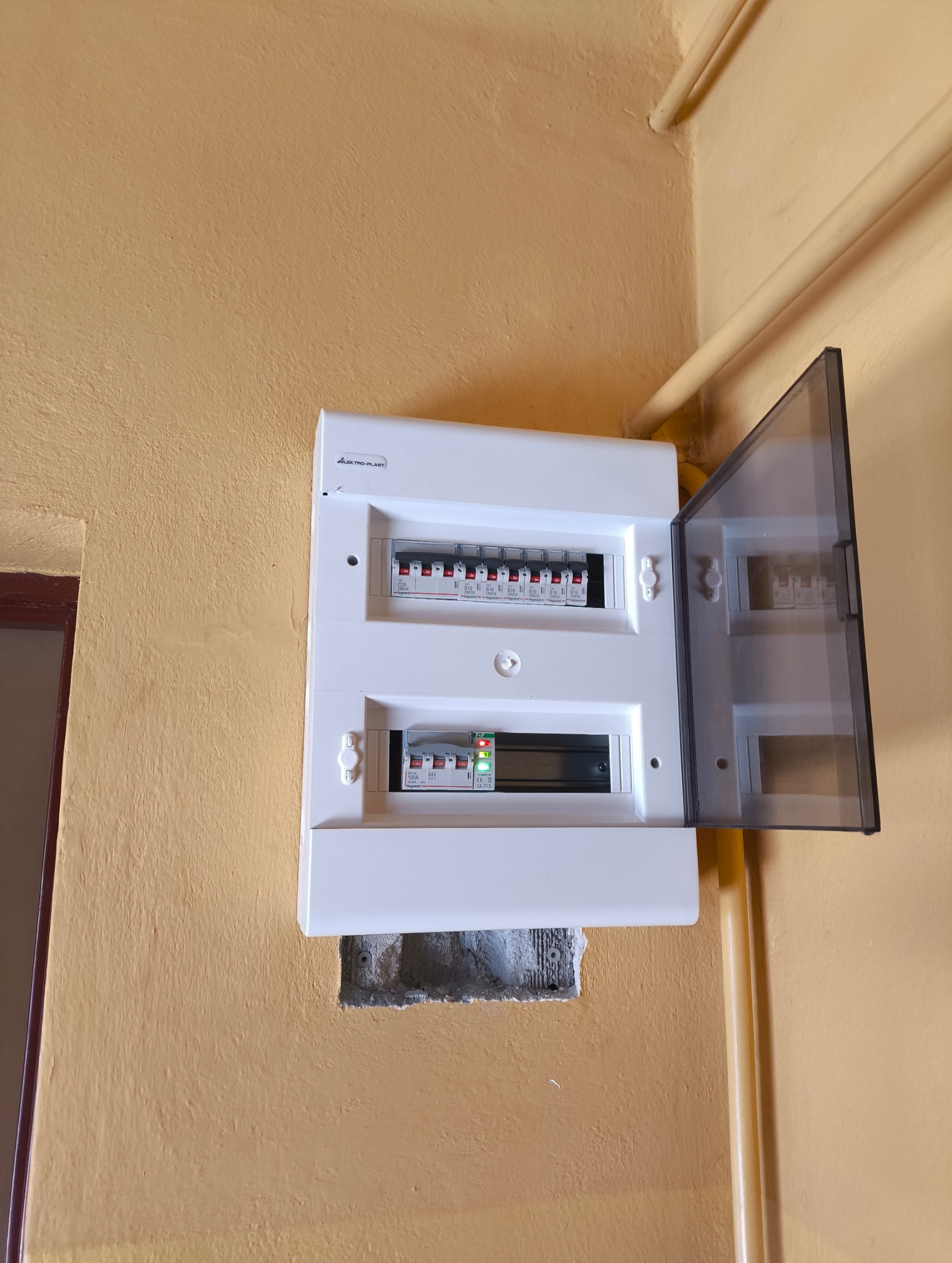 Biała skrzynka elektryczna marki Elektro-Plast zamontowana na żółtej ścianie, z widocznymi bezpiecznikami i otwartą, przezroczystą klapką.