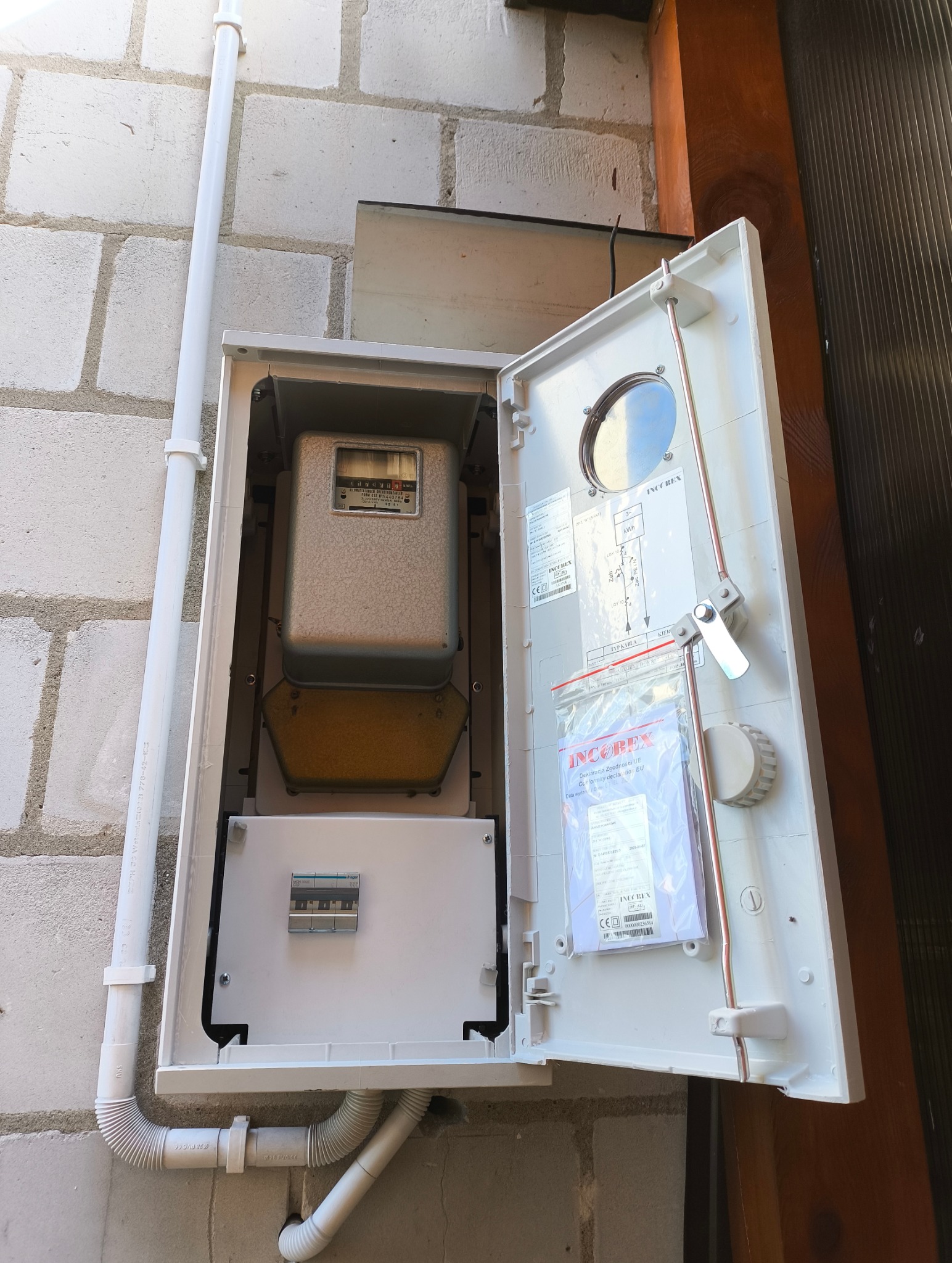 Otwarta skrzynka z licznikiem elektrycznym na szarej ścianie z bloczków. Widoczne przewody, schemat i dokumenty wewnątrz. Detal instalacji.