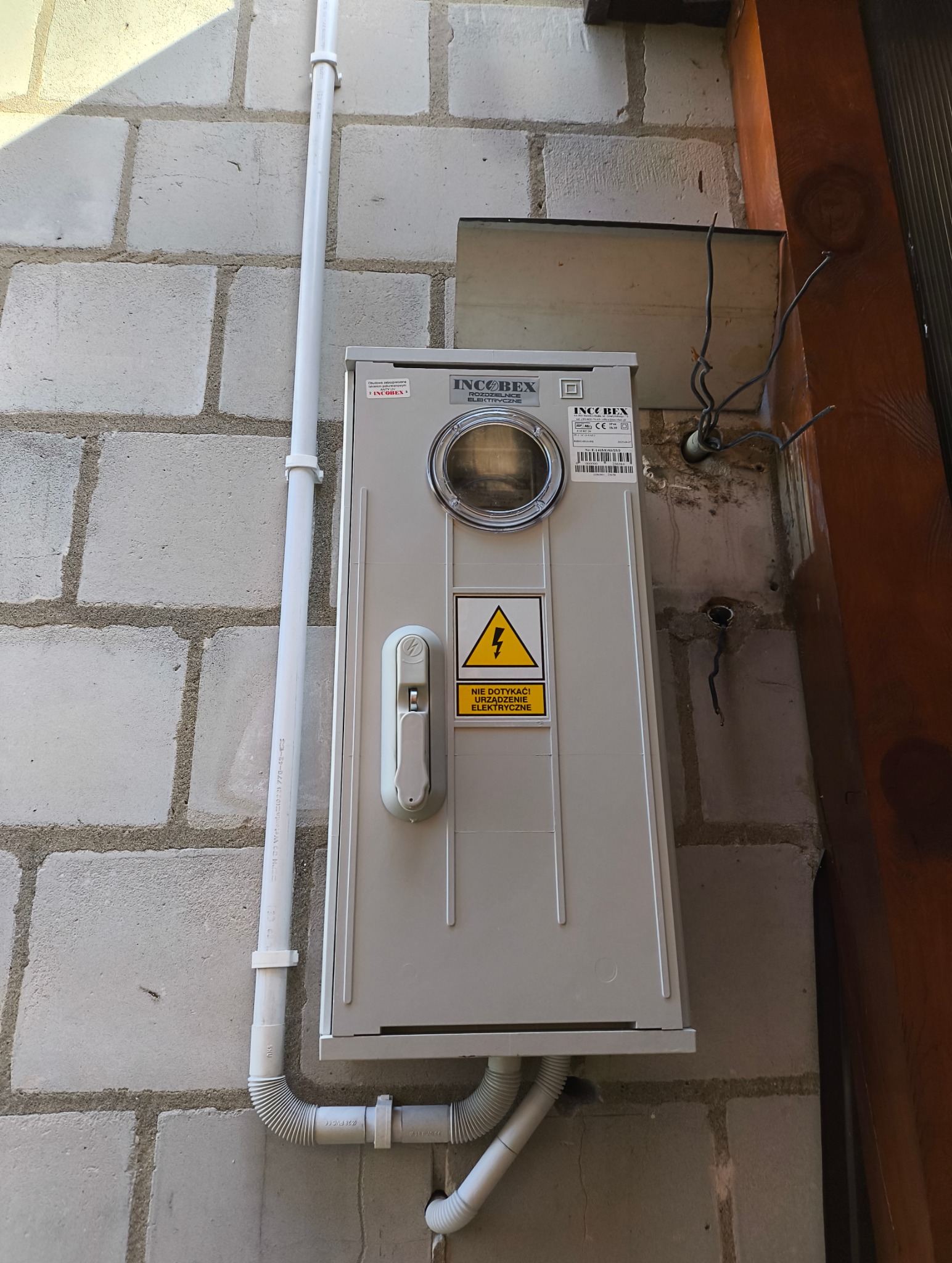 Szara skrzynka z licznikiem elektrycznym i ostrzeżeniem 'Nie dotykać! Urządzenie elektryczne' zamontowana na szarej ścianie z betonowych pustaków.