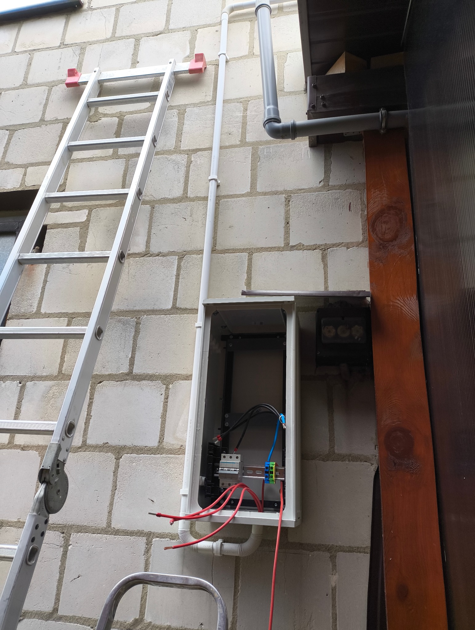 Otwarta skrzynka elektryczna na ścianie z bloków, widoczne przewody, rurki PCV, obok drabina oparta o ścianę. Element instalacji elektrycznej w trakcie prac.