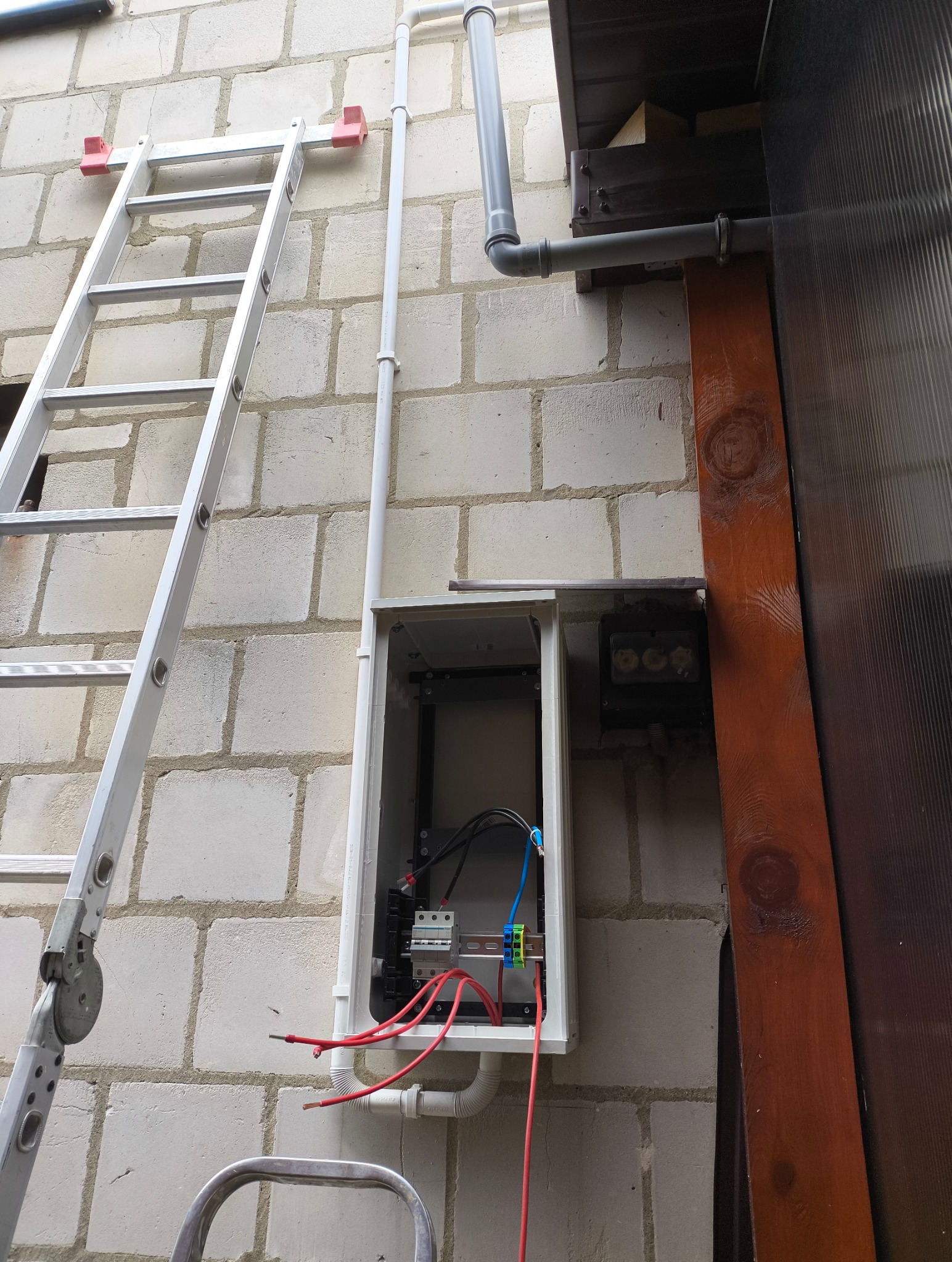 Skrzynka elektryczna z przewodami na ścianie z bloków, obok drabina i rury. Widoczne elementy instalacji elektrycznej w trakcie montażu.
