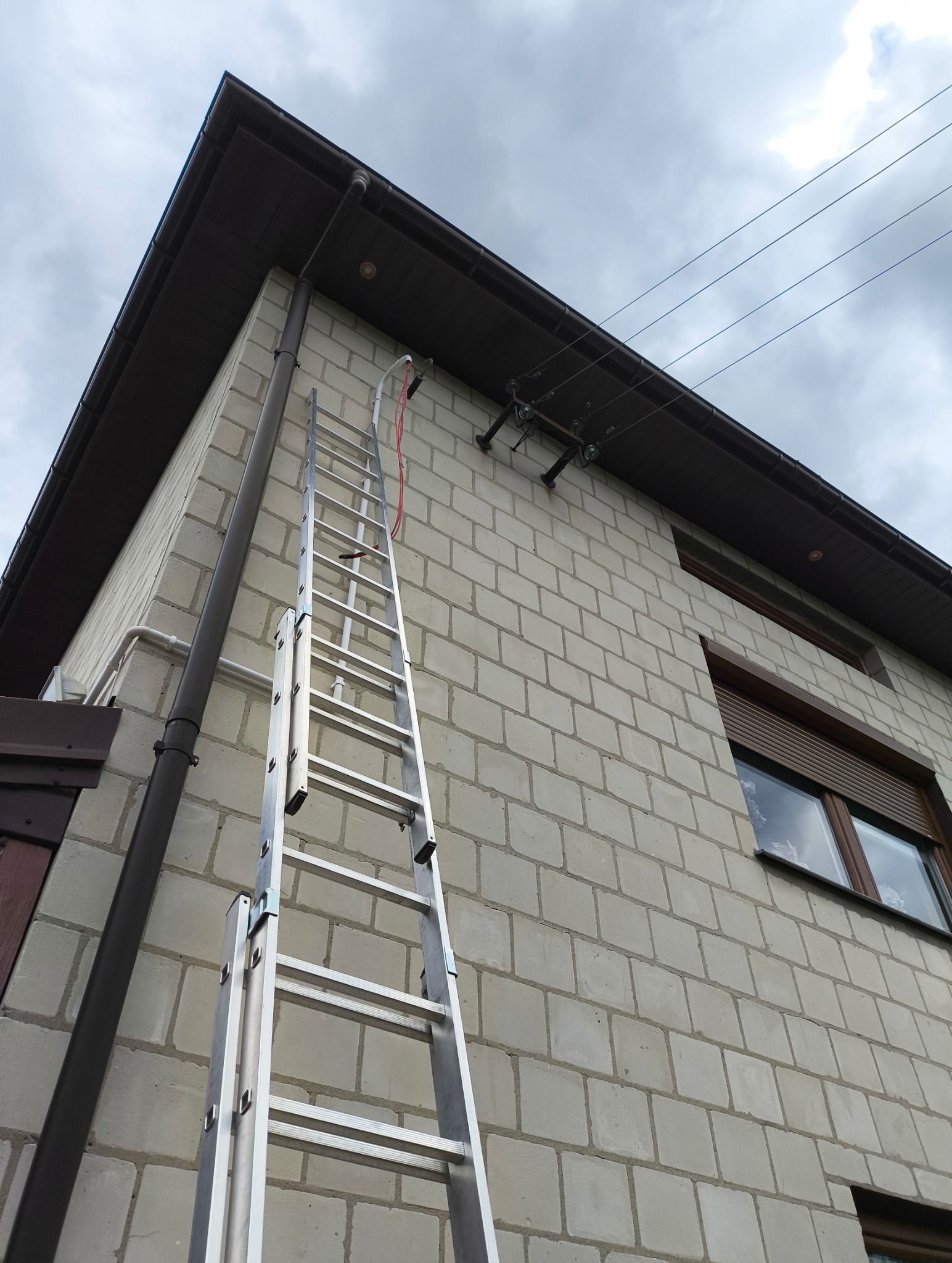 Aluminiowa drabina oparta o ścianę budynku z instalacją elektryczną na zewnątrz, widok z dołu, pochmurne niebo w tle. Czerwony przewód wzdłuż drabiny.