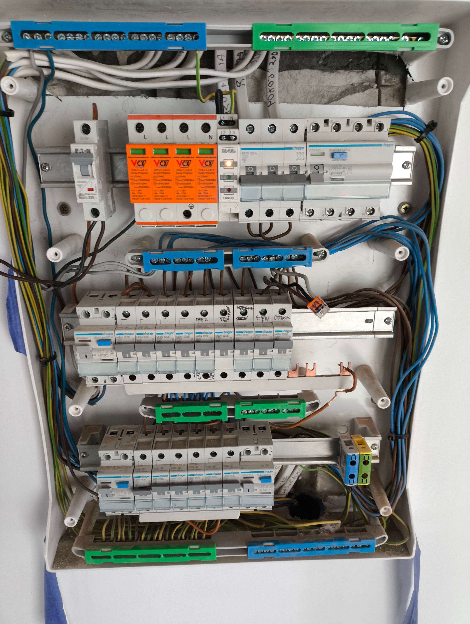 Rozdzielnica elektryczna z zabezpieczeniami, widoczne kable, bezpieczniki i ograniczniki przepięć. Instalacja w białej obudowie, z elementami w kolorach: niebieski, zielony, pomarańczowy.