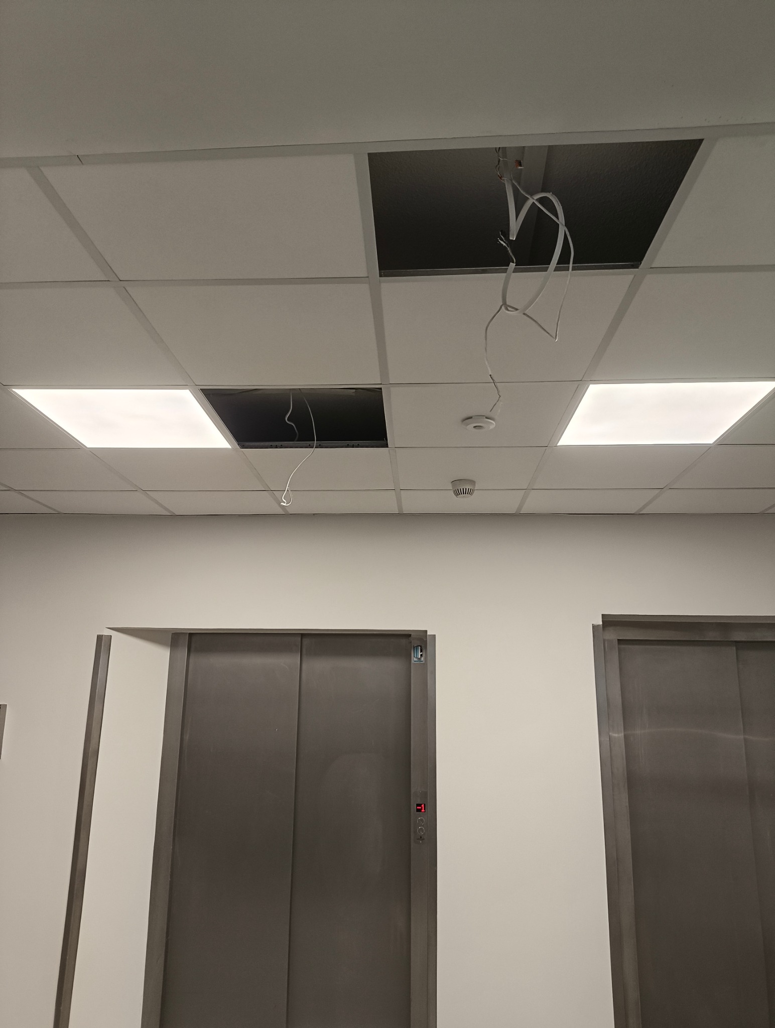 Otwarty panel sufitowy z wiszącymi kablami w biurze, widoczne windy. Prace elektryczne w trakcie, instalacja oświetlenia lub czujników.