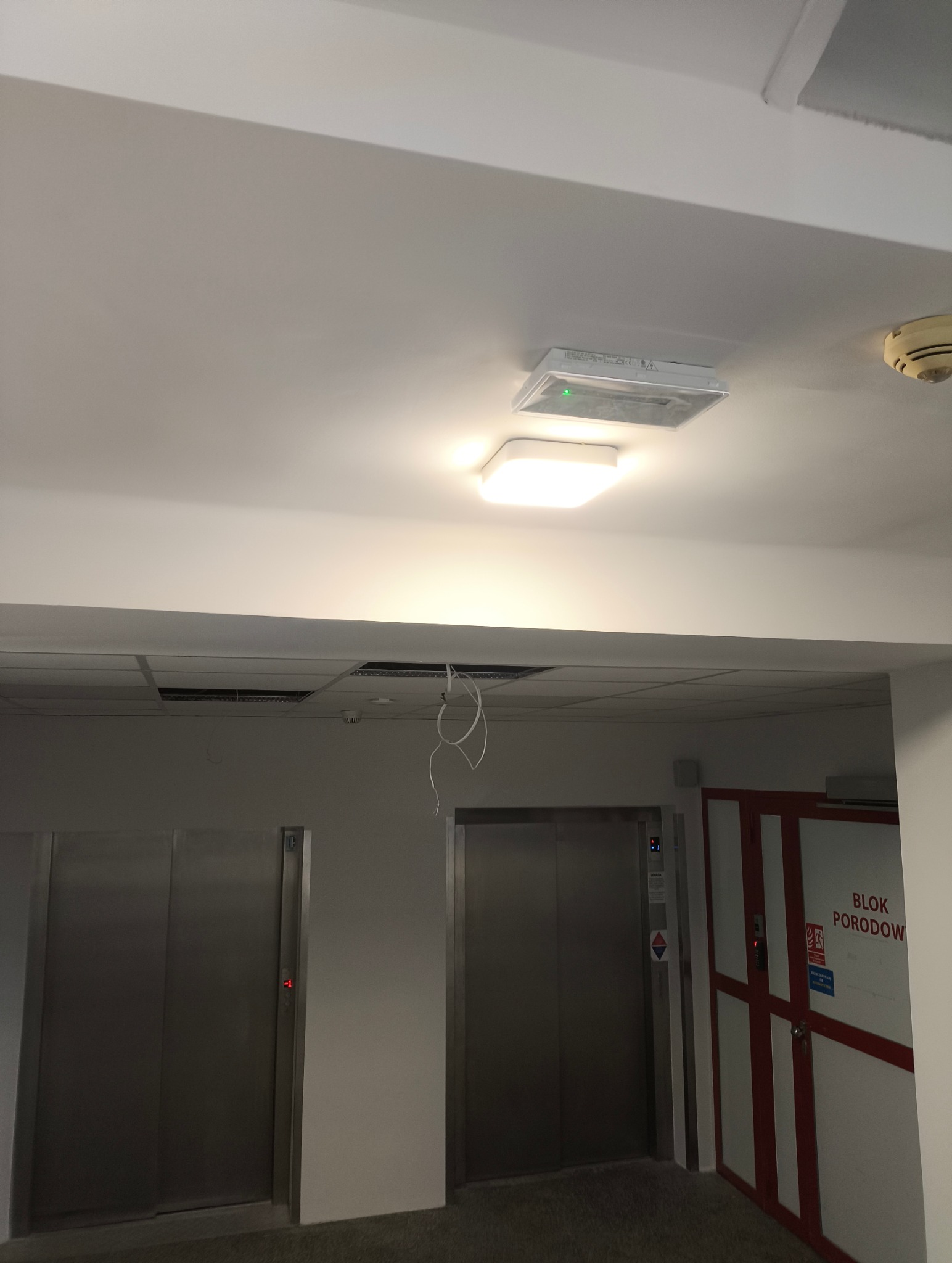 Nowoczesne oświetlenie sufitowe LED w korytarzu budynku z windami, widoczne przewody i czujnik dymu. Jasne światło podkreśla minimalistyczny design.