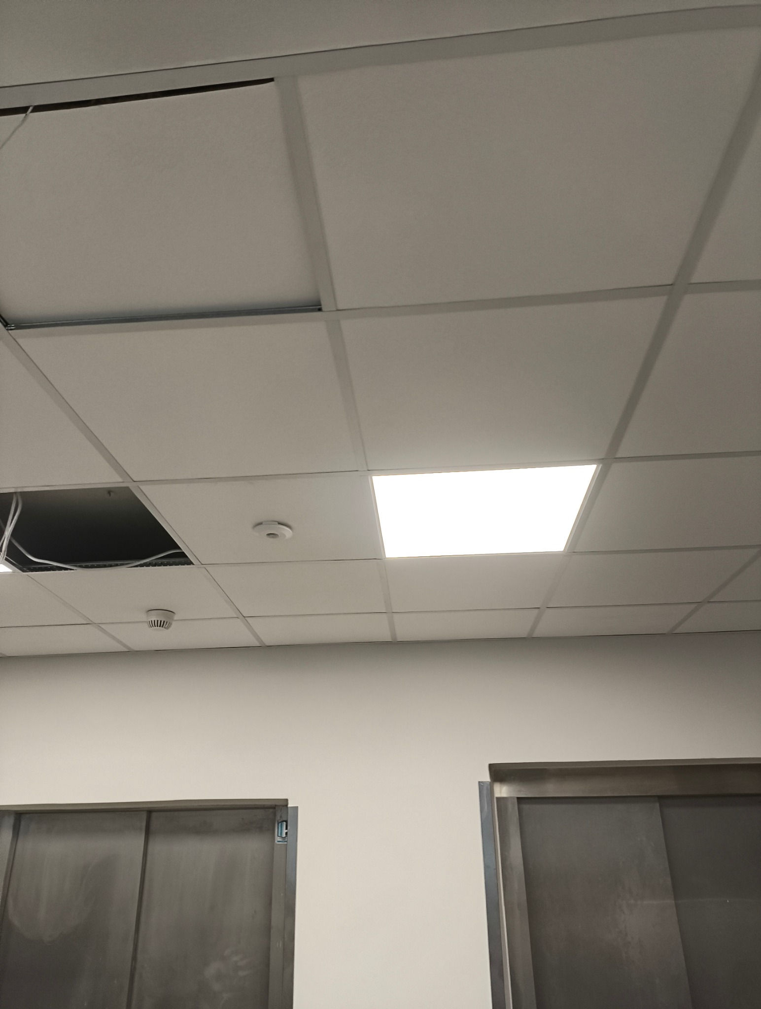 Biały sufit podwieszany z widocznymi kablami i czujnikiem dymu. Jedna z płyt sufitowych jest zdjęta, odsłaniając instalację.