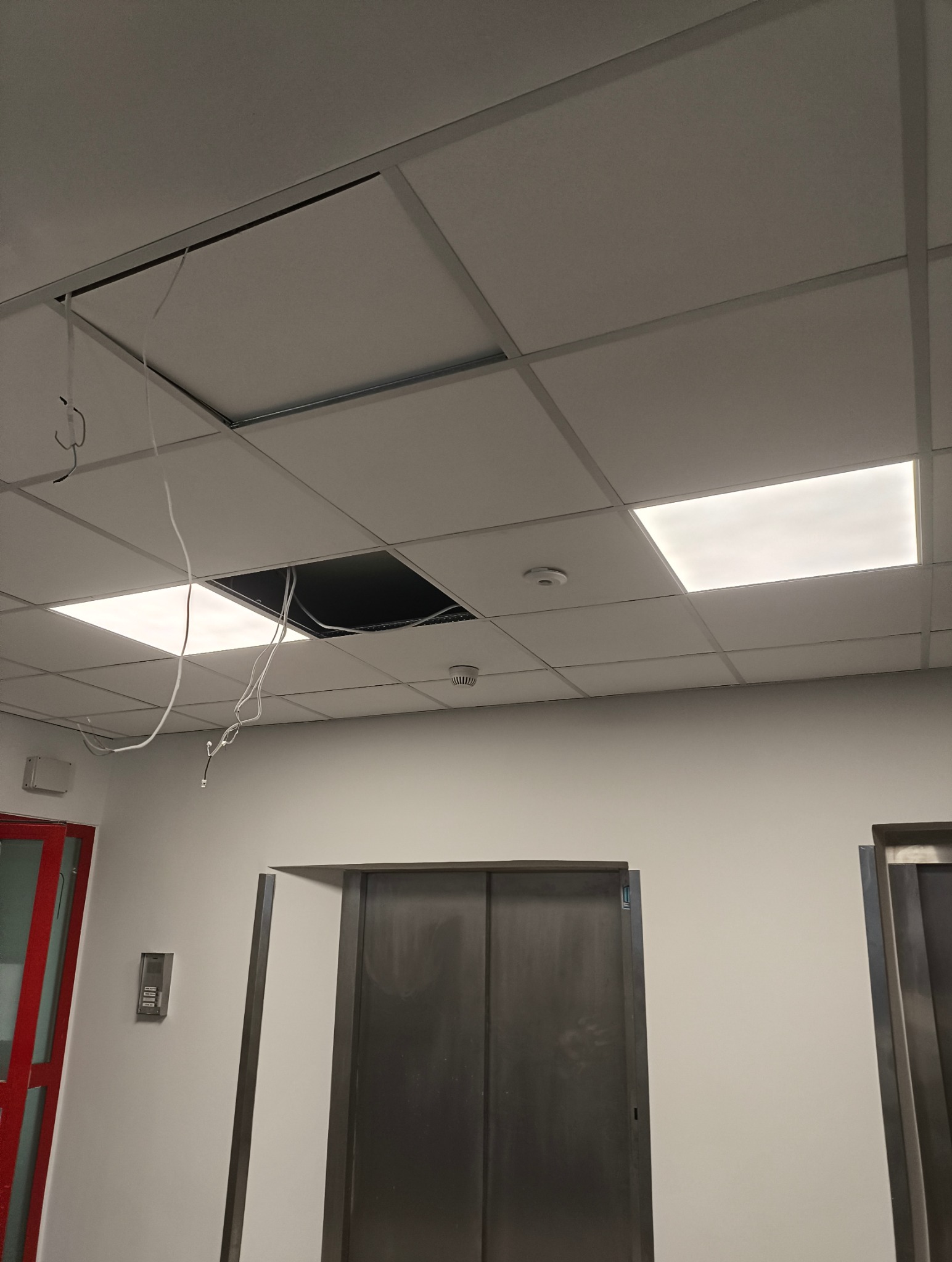 Kasetonowy sufit podwieszany z brakującym panelem i wystającymi przewodami. Obok widoczny panel oświetleniowy i czujnik dymu.