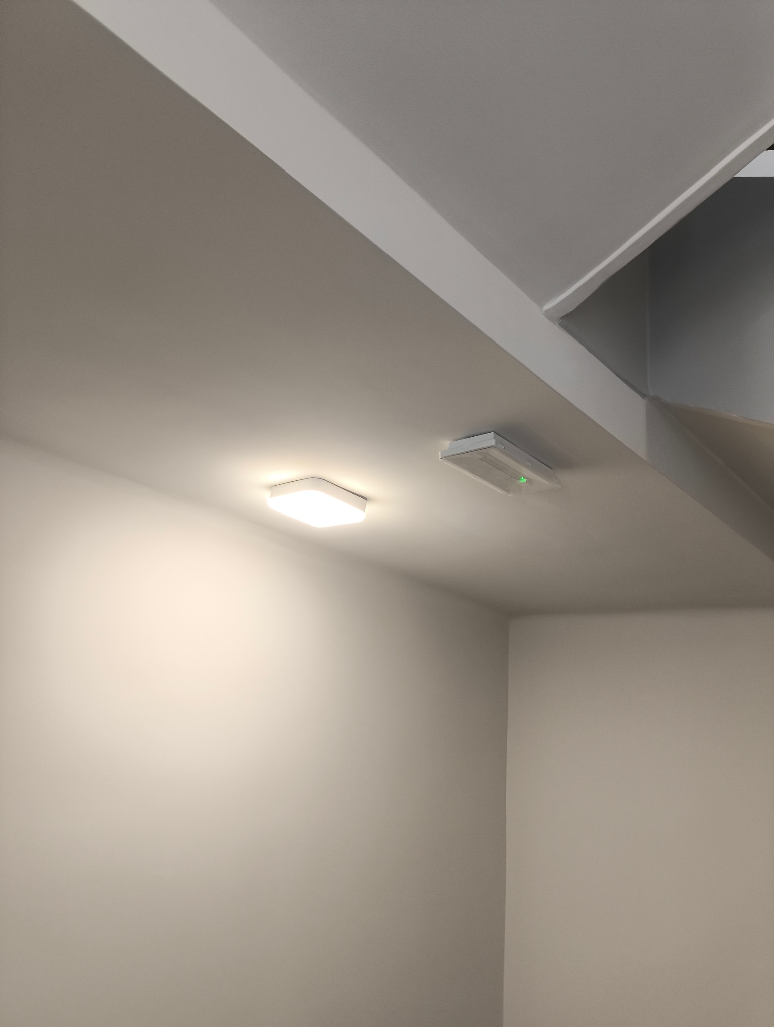 Zamontowane, nowoczesne oświetlenie sufitowe w minimalistycznym wnętrzu. Widoczna lampa LED i czujnik dymu z zieloną diodą. Neutralna kolorystyka ścian.