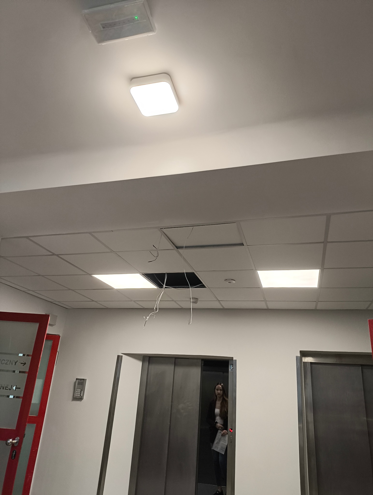 Sufit podwieszany z otwartym panelem, widoczne przewody. W tle osoba w windzie. Oświetlenie sufitowe. Prace remontowe lub naprawa. Widok z dołu.