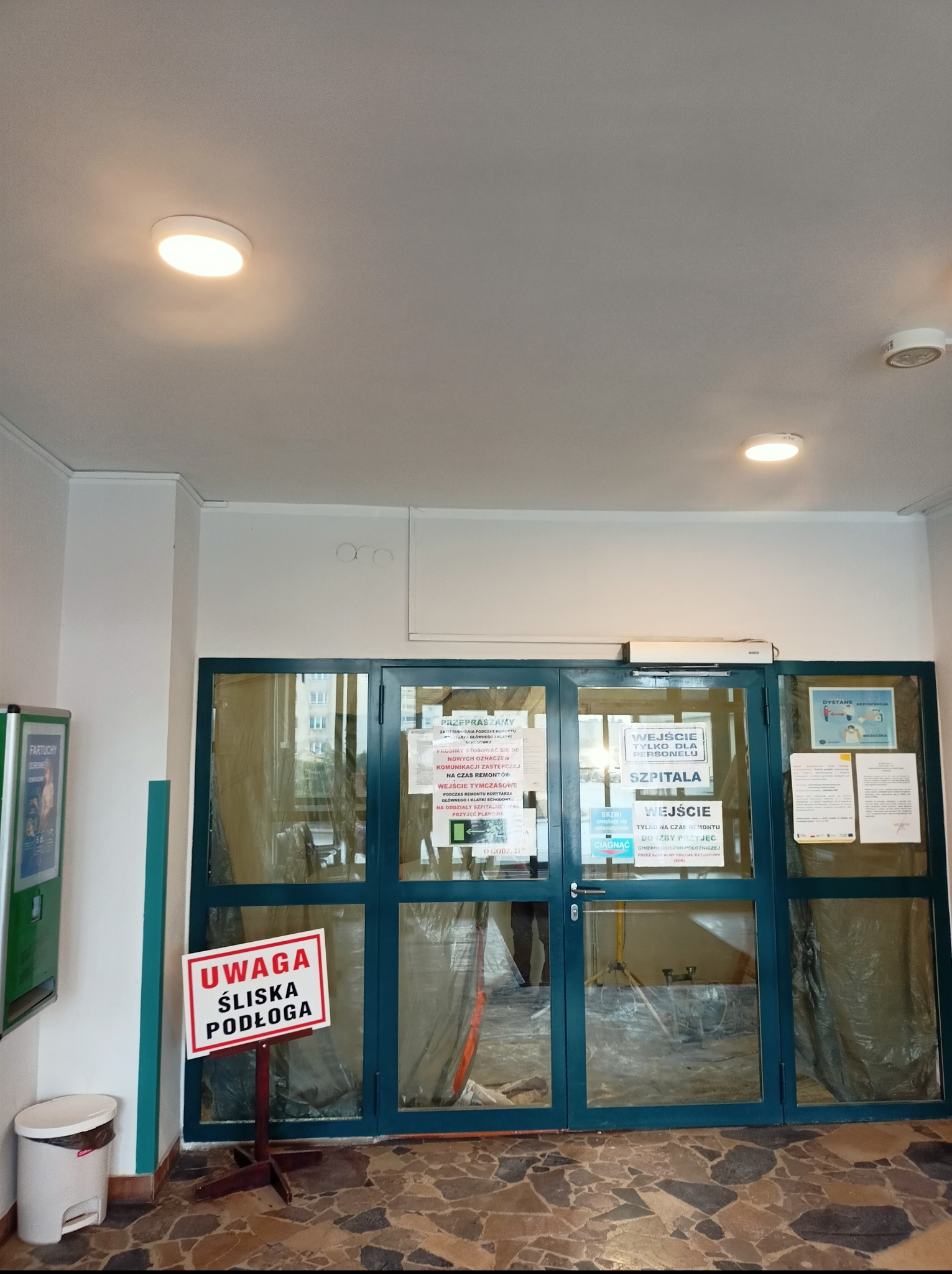 Okrągłe lampy sufitowe w szpitalnym korytarzu z remontem w tle, widoczne przez przeszklone drzwi. Tabliczka ostrzegawcza o śliskiej podłodze.
