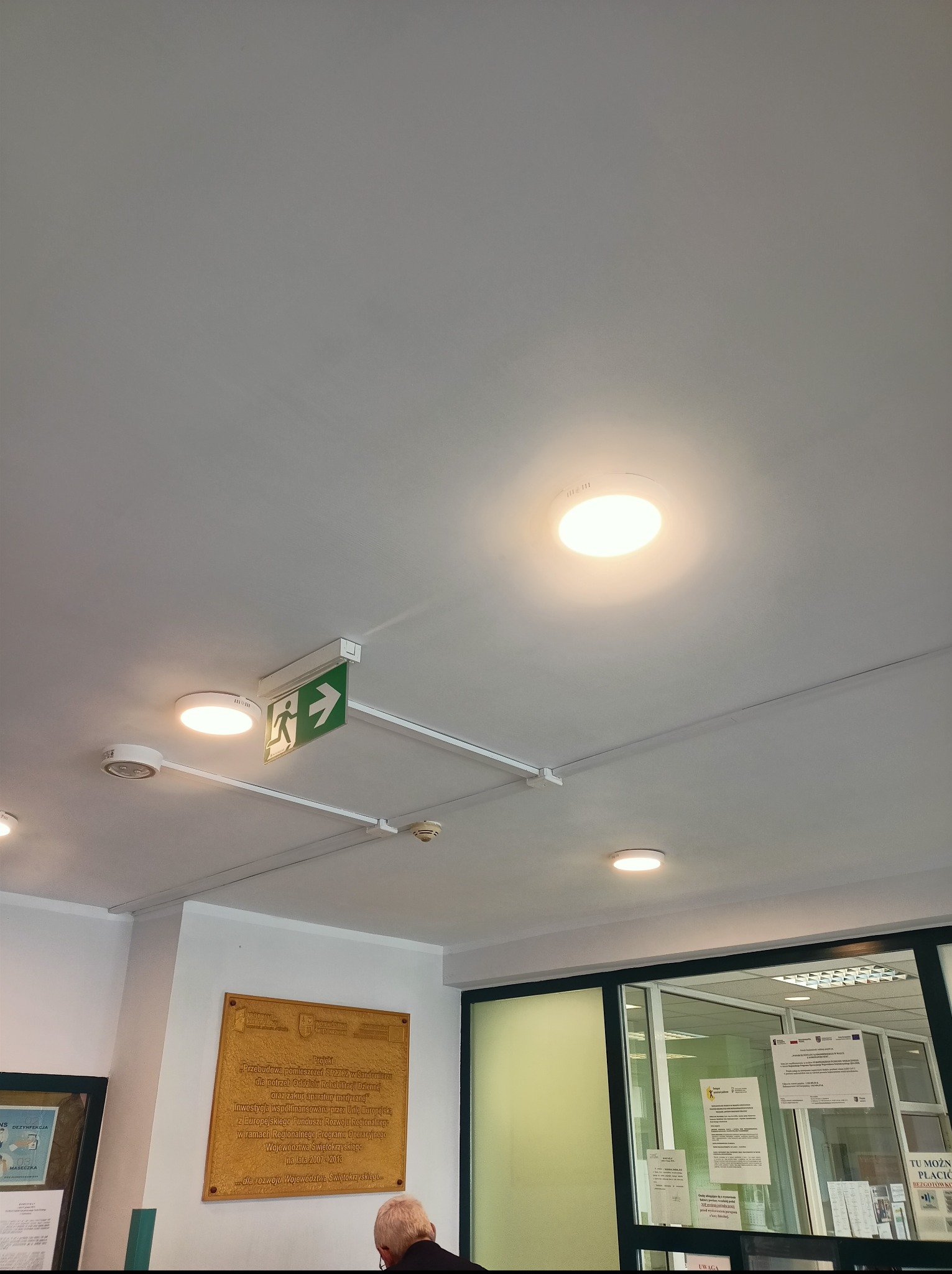 Biały sufit z zamontowanymi lampami LED i zielonym znakiem ewakuacyjnym z piktogramem biegnącej osoby. Widoczne przewody i czujnik dymu. Ujęcie z dołu.