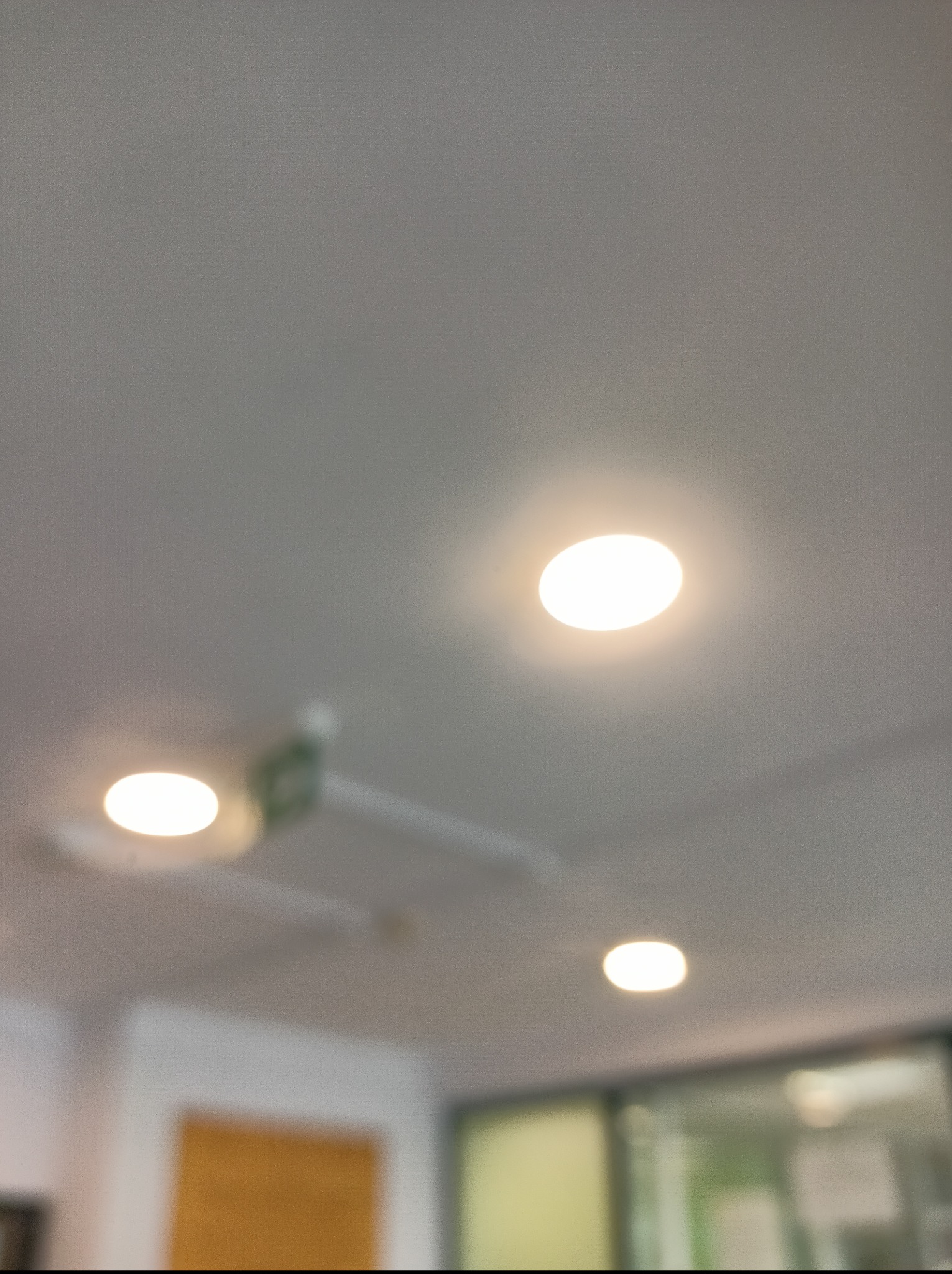 Biały sufit z trzema okrągłymi, wpuszczanymi lampami LED. Jedna z lamp częściowo zasłonięta przez element wentylacyjny. Rozproszone światło.