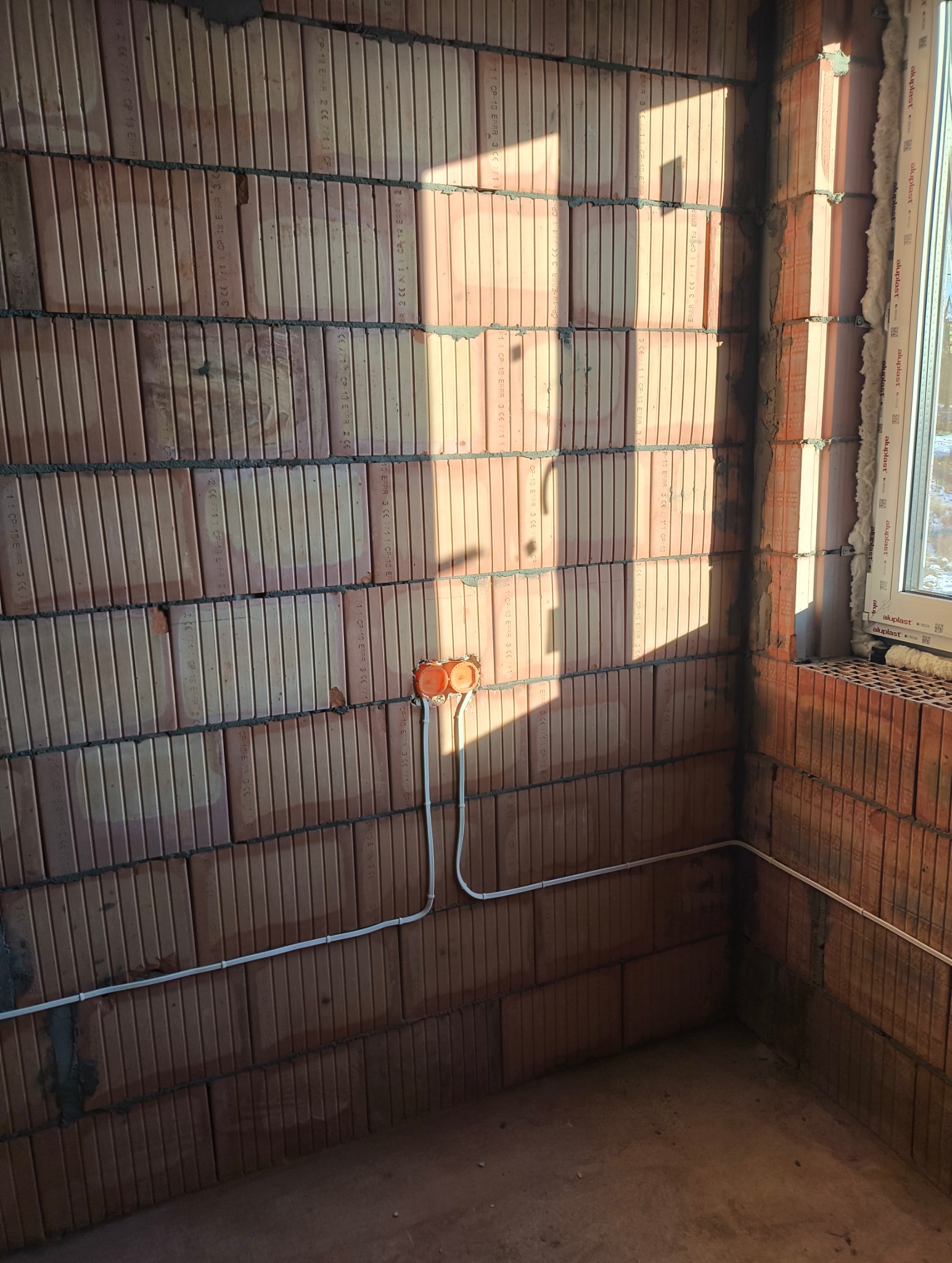 Wewnętrzna instalacja elektryczna w trakcie montażu: pomarańczowa puszka z przewodami na ścianie z pustaków ceramicznych, widoczne przewody. Oświetlenie słoneczne.