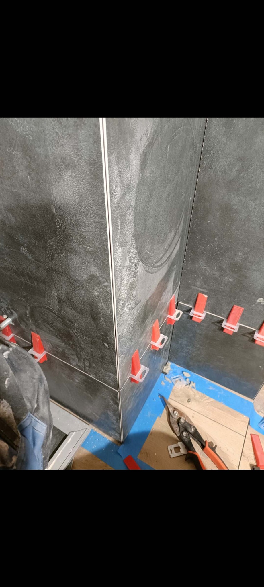 Układanie ciemnych płytek w rogu pomieszczenia z użyciem czerwonych klipsów poziomujących. Widoczne narzędzia glazurnicze i fragment podłogi zabezpieczonej taśmą.