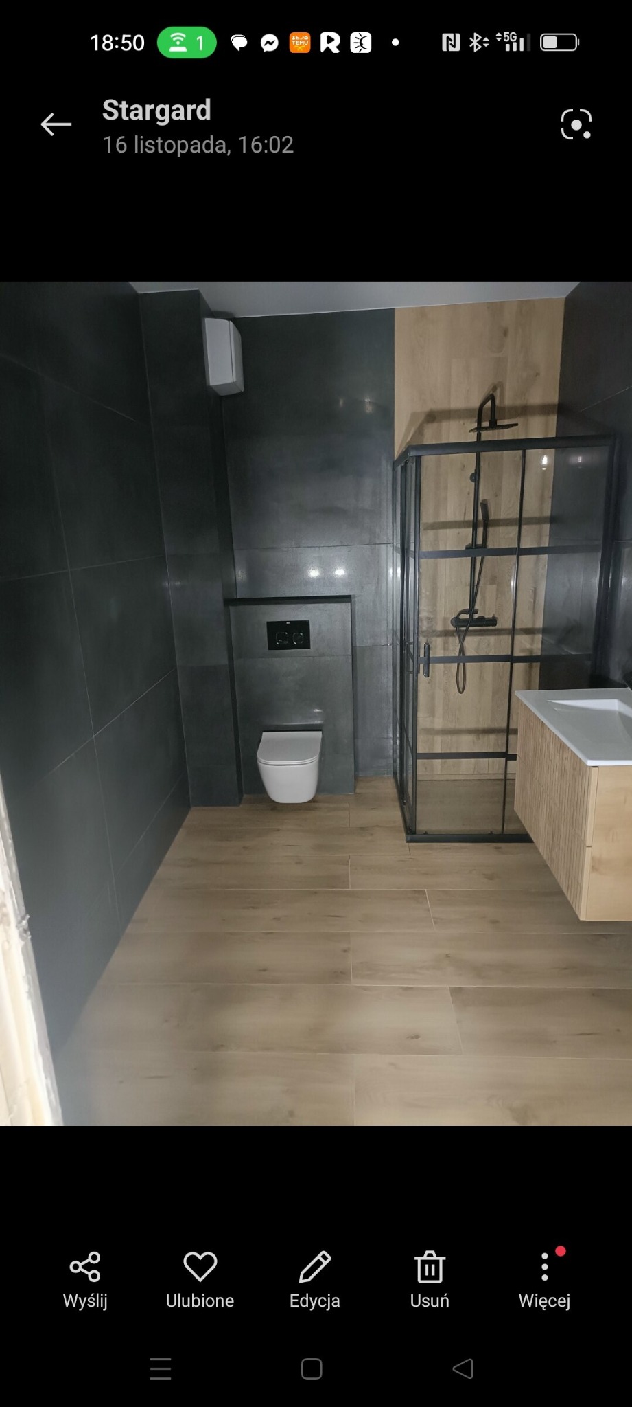 Nowoczesna łazienka po remoncie: szare płytki ścienne, drewnopodobna podłoga, kabina prysznicowa typu walk-in z czarnymi ramami i wisząca toaleta.