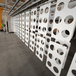 Lakiernia Proszkowa Raltom - Białe, perforowane panele zawieszone na linii produkcyjnej po malowaniu proszkowym. Widoczne elementy konstrukcyjne i pracownik w tle.