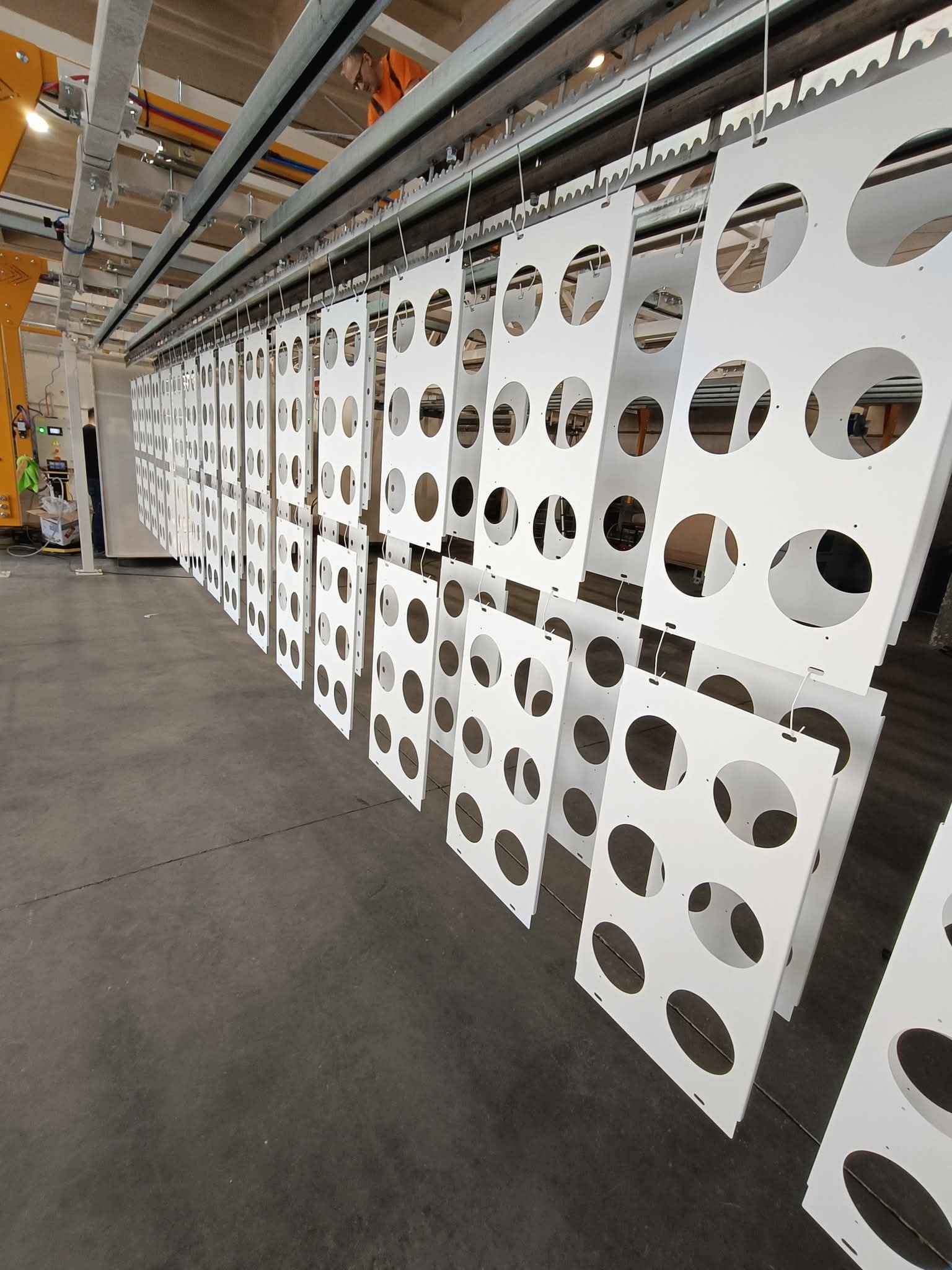 Białe, perforowane panele zawieszone na linii produkcyjnej po malowaniu proszkowym. Widoczne elementy konstrukcyjne i pracownik w tle.