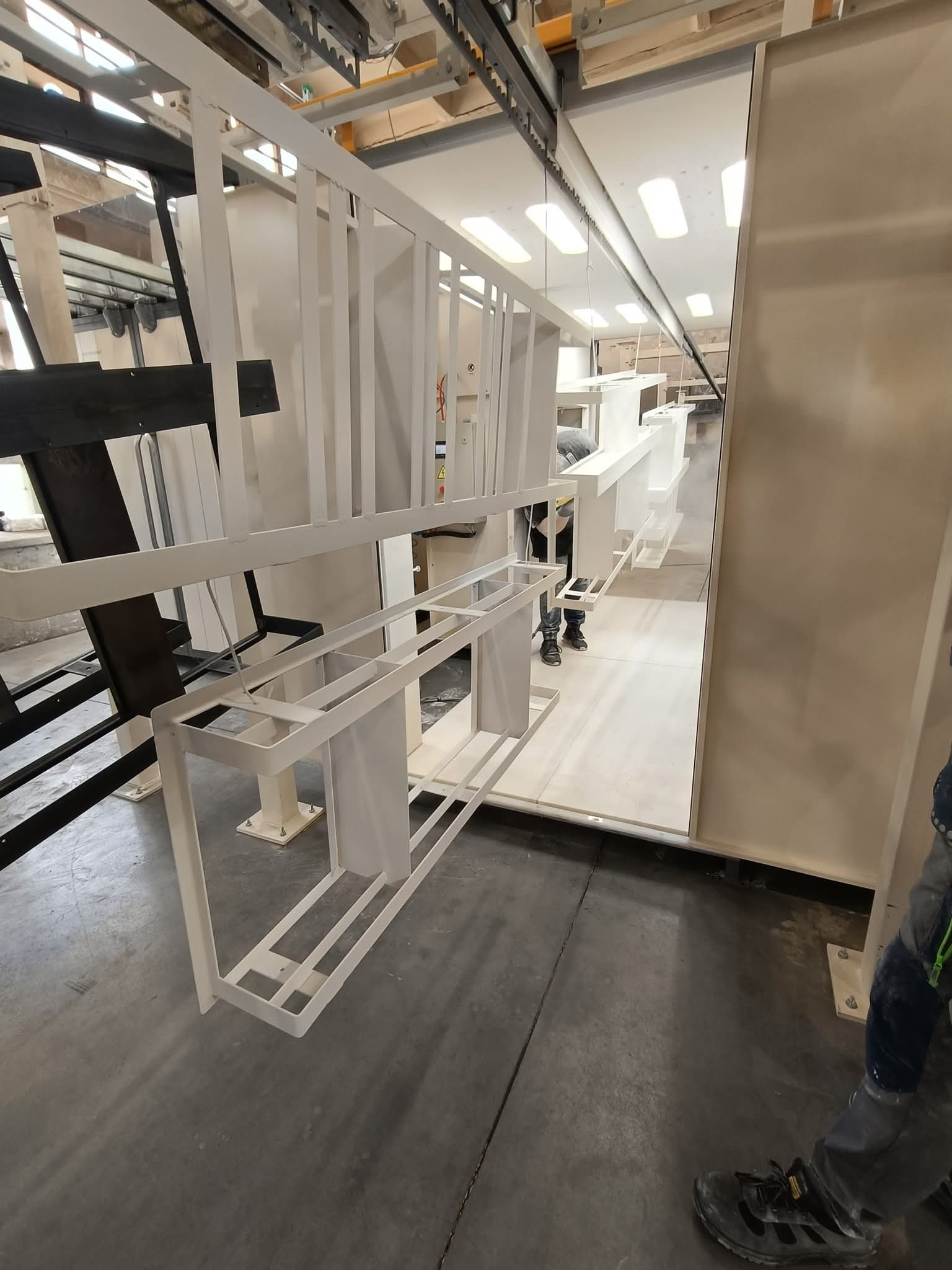 Elementy konstrukcyjne po nałożeniu białej farby, zawieszone w hali produkcyjnej. Widoczna część linii technologicznej i pracownik w tle. Detal wykończenia.