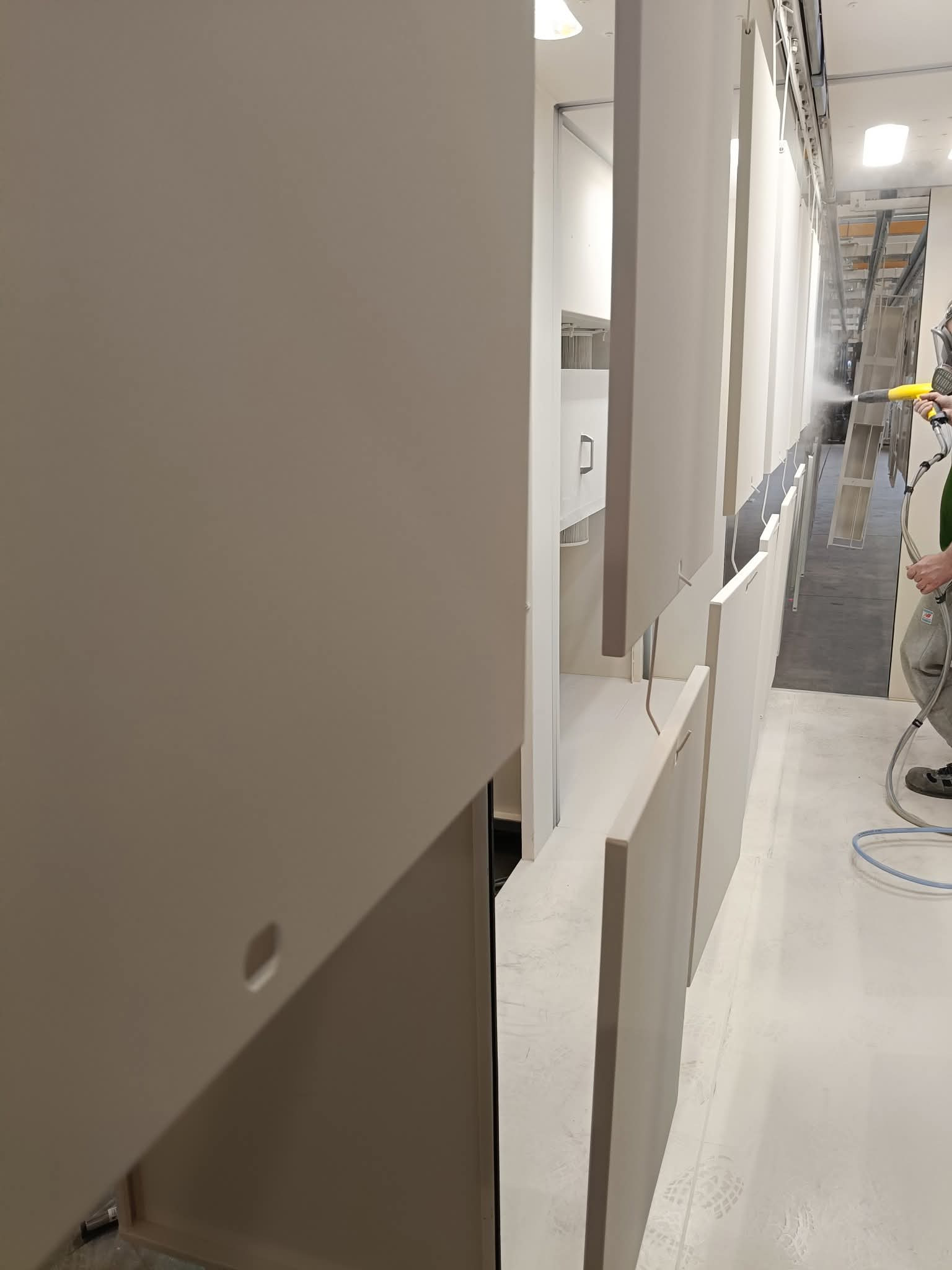 Lakierowanie frontów meblowych w kabinie lakierniczej. Widoczny pracownik w masce ochronnej i z pistoletem natryskowym, aplikujący farbę na zawieszone elementy.