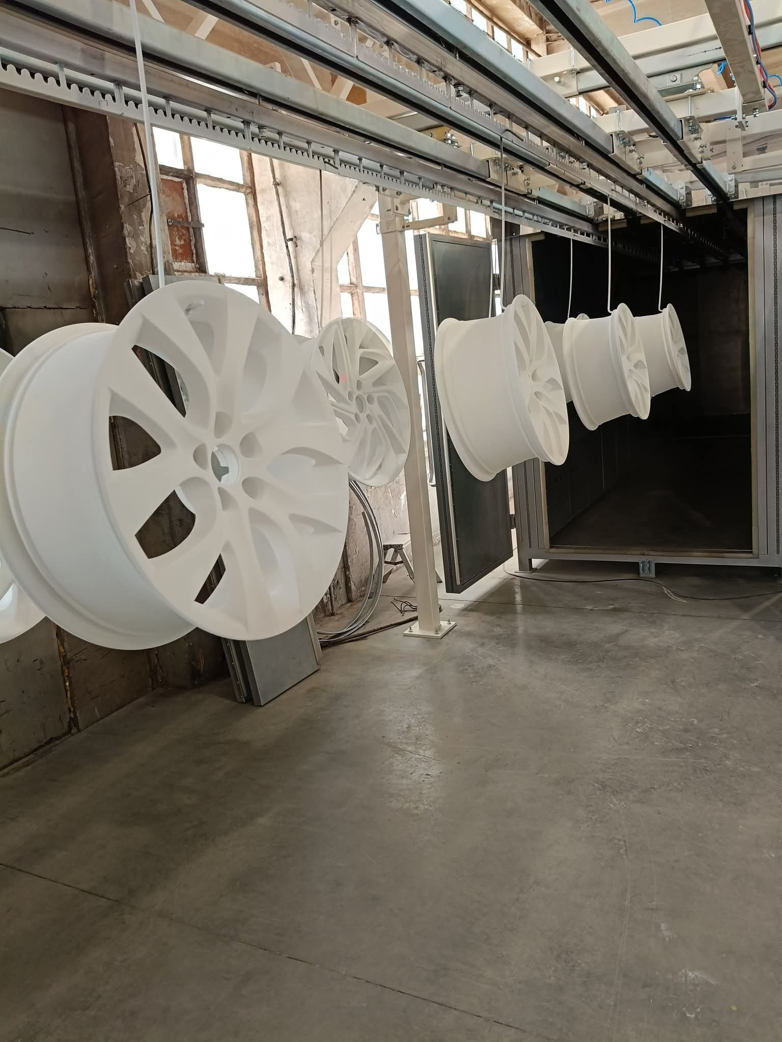 Białe felgi aluminiowe po obróbce, zawieszone na szynie w hali przemysłowej. Widoczny piec do wygrzewania.