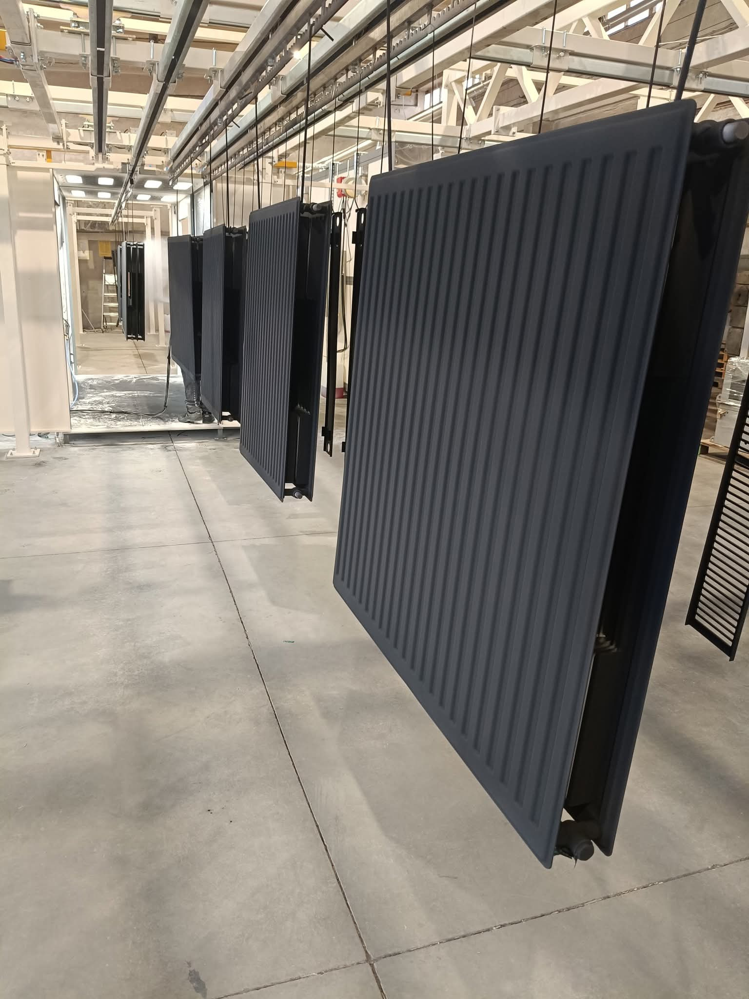 Szare, wiszące grzejniki w hali, po obróbce. Widoczne detale konstrukcji nośnej i oświetlenie przemysłowe. Grzejniki równomiernie rozmieszczone.