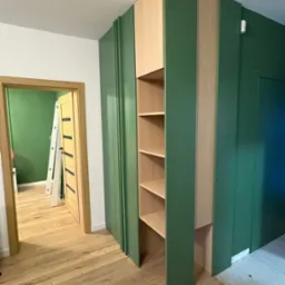 Zielona zabudowa meblowa z półkami z jasnego drewna, w tle widoczne drzwi i drabina. Podłoga z paneli imitujących drewno. Nietypowe połączenie kolorów.