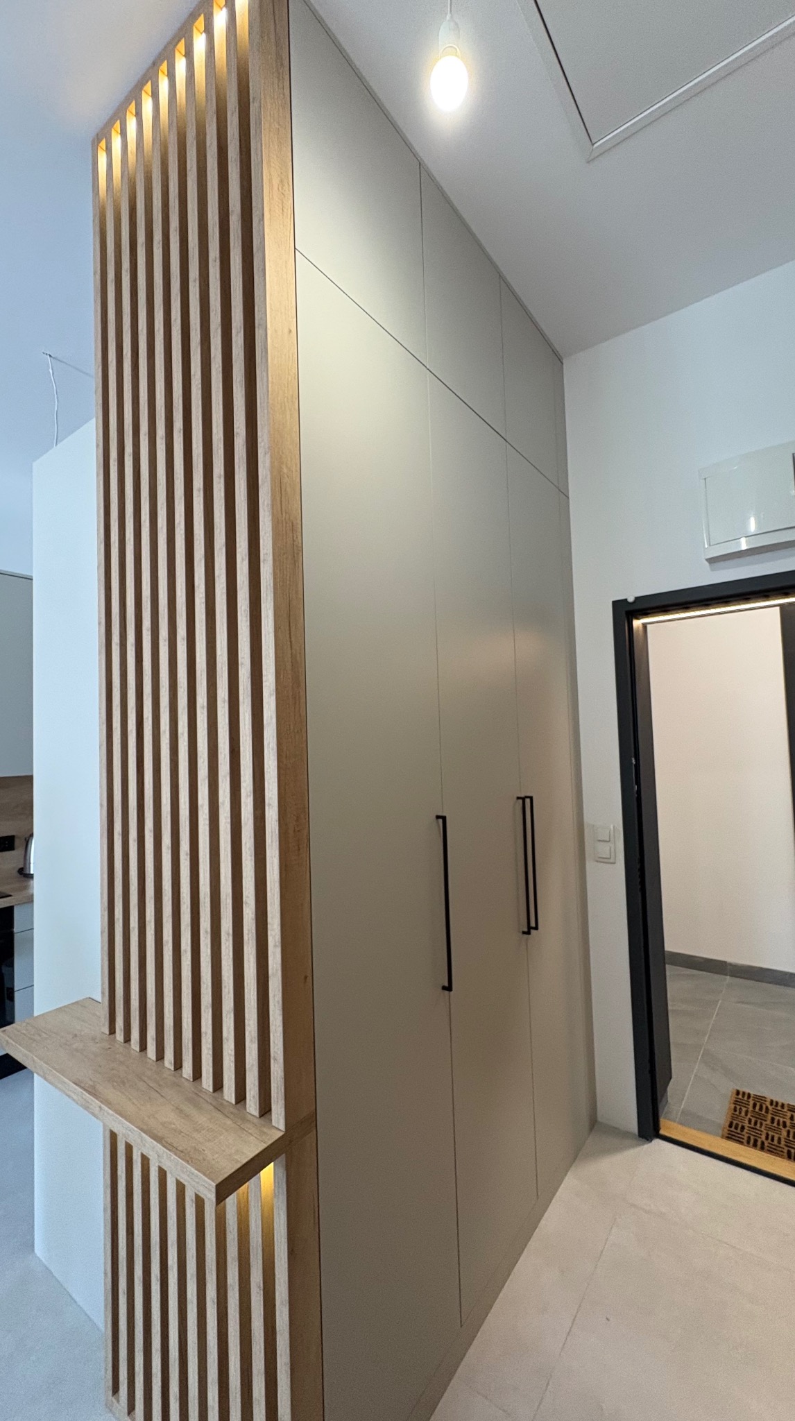 Nowoczesna szafa w zabudowie z drewnianą ażurową ścianką działową i półką, podświetlana od góry. Minimalistyczne czarne uchwyty i oświetlenie sufitowe.