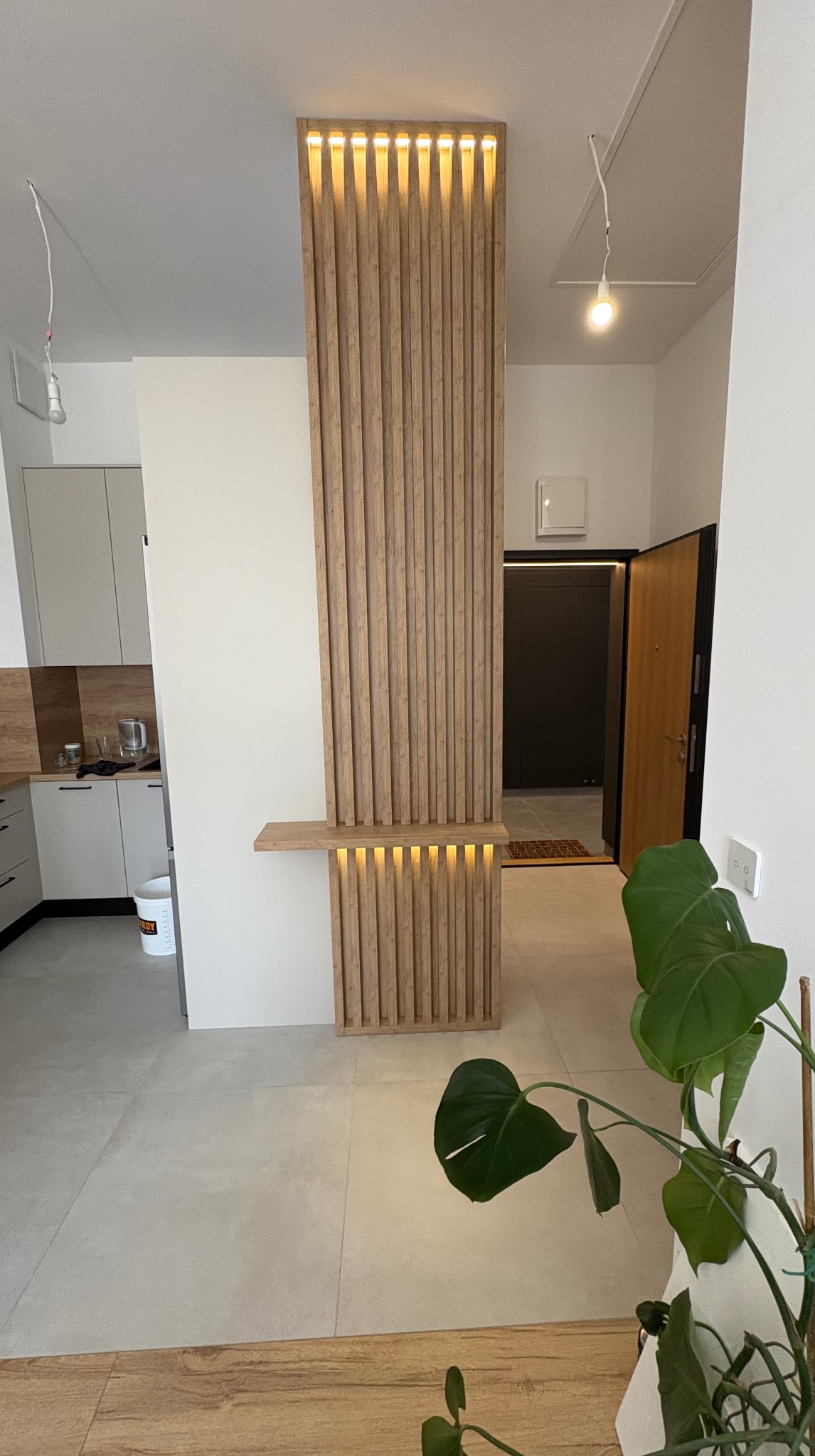 Drewniana ścianka działowa z podświetleniem LED oddziela kuchnię od przedpokoju. Nowoczesny design z elementami naturalnego drewna i minimalistycznym oświetleniem.