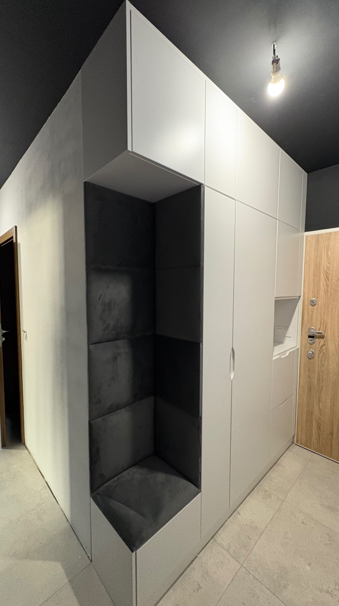 Nowoczesna zabudowa meblowa w przedpokoju z siedziskiem tapicerowanym w kolorze grafitowym. Minimalistyczny design, szare ściany i podłoga.