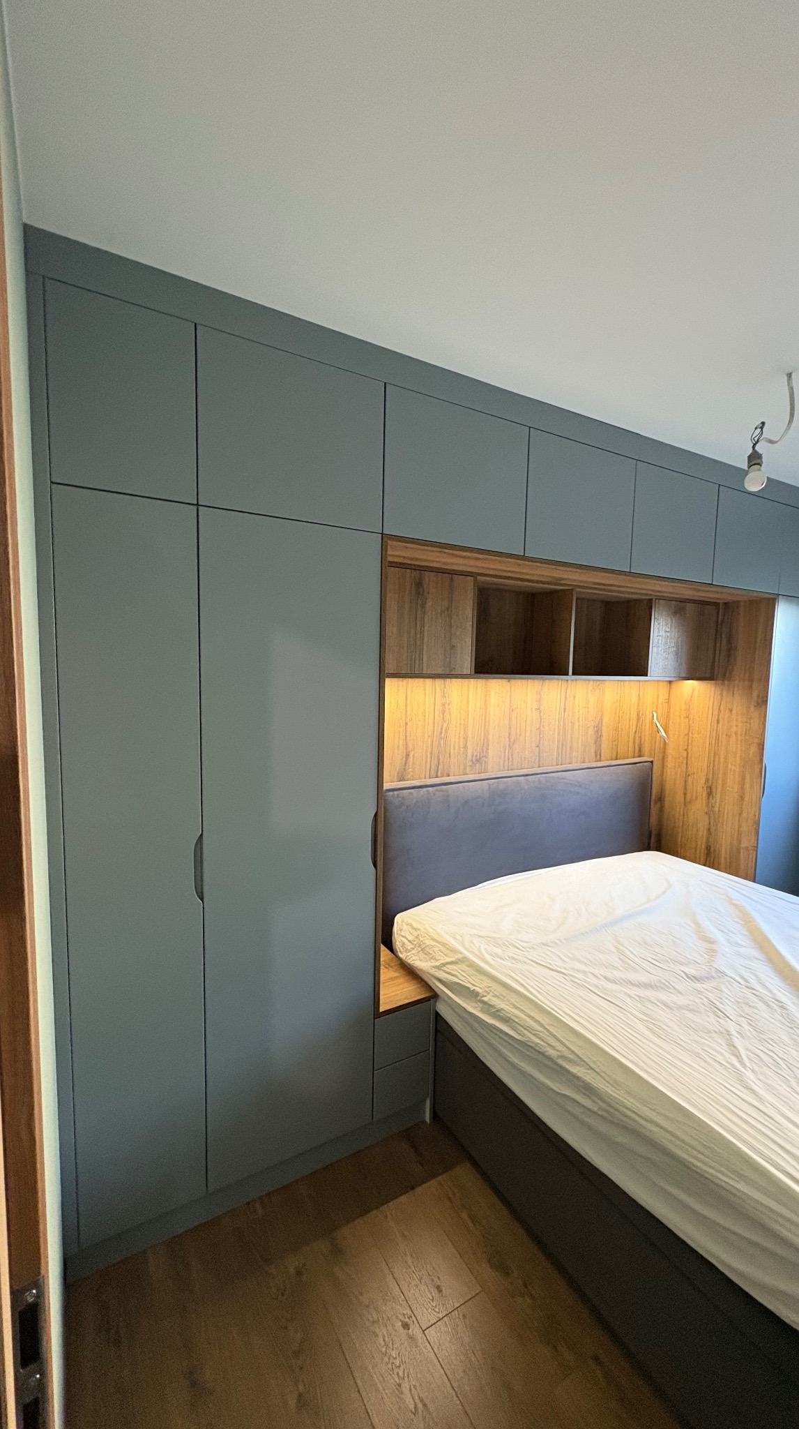 Szara zabudowa sypialni z szafami i wnęką z drewnianym wykończeniem nad łóżkiem. Oświetlenie wnęki podkreśla ciepły odcień drewna. Widoczna podłoga z paneli.