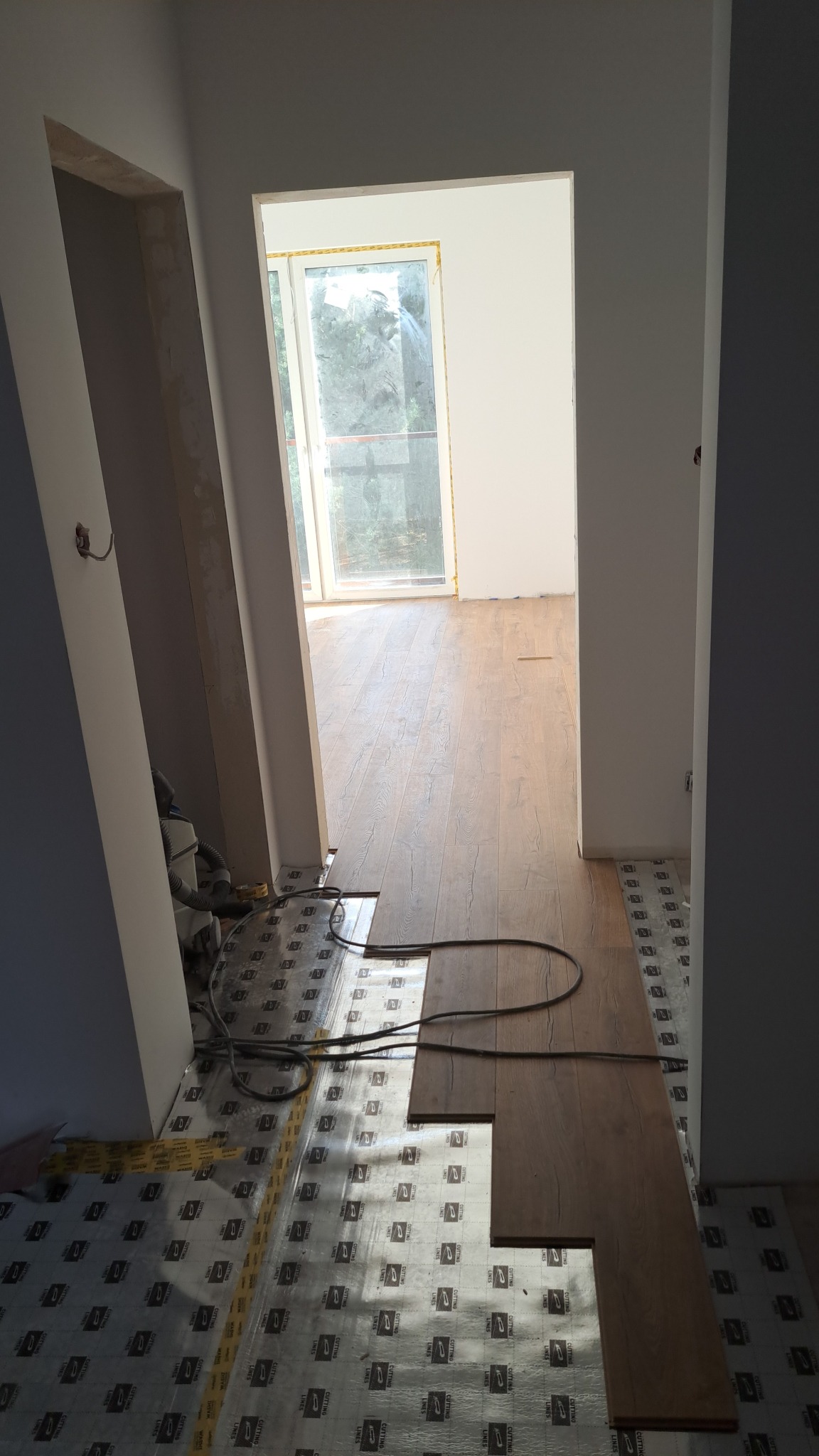 Układanie paneli podłogowych w korytarzu. Widoczne panele z wzorem drewna, folia izolacyjna i odkurzacz przemysłowy. Częściowo wykończone ściany i futryny.