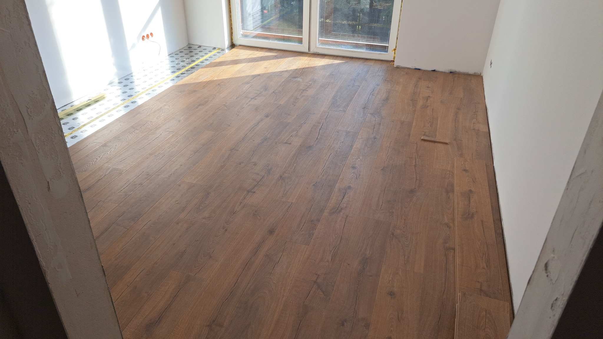 Układanie paneli podłogowych w jasnym pokoju z widokiem na okno balkonowe. Widoczna podkładka pod panele. Częściowo ułożona podłoga z paneli o wzorze drewna.