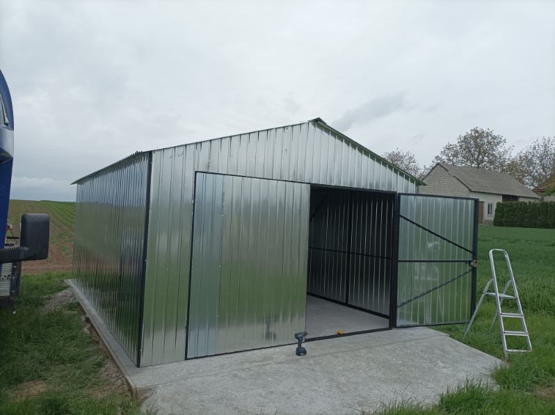 Błyszczący, stalowy garaż blaszany z otwartymi dwuskrzydłowymi drzwiami, widoczna drabina oparta o ścianę, na betonowej wylewce, w tle budynek mieszkalny.