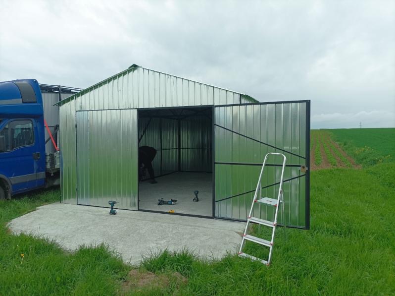 Montaż garażu blaszanego na betonowej wylewce. Otwarta brama, w tle drabina i pracownik. Widoczne narzędzia. Obok zaparkowany samochód dostawczy i pole uprawne.