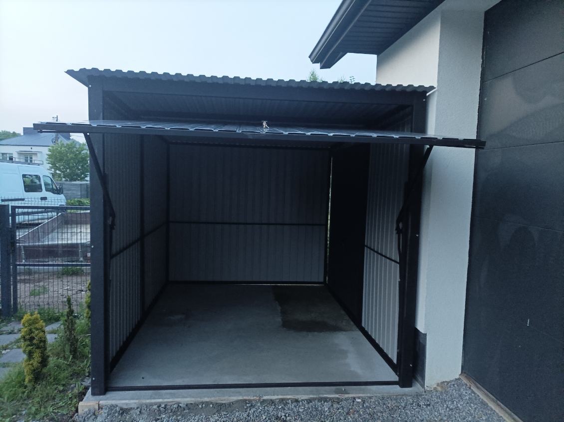 Otwarty garaż blaszany z metalowym dachem, przylegający do budynku z szarą elewacją. Widoczny fragment podjazdu i ogrodzenia.
