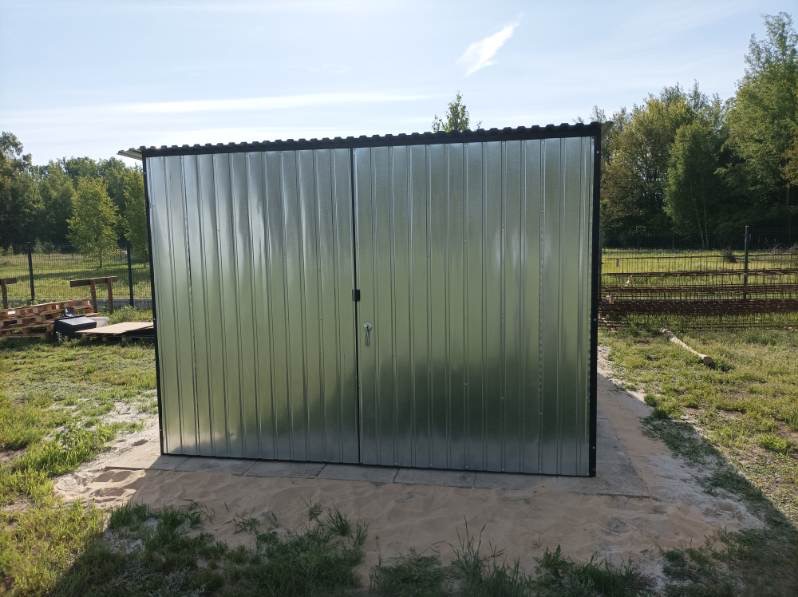 Gotowy garaż blaszany z dwuskrzydłowymi drzwiami, ustawiony na betonowej wylewce na tle zieleni. Widoczne palety i elementy konstrukcyjne.