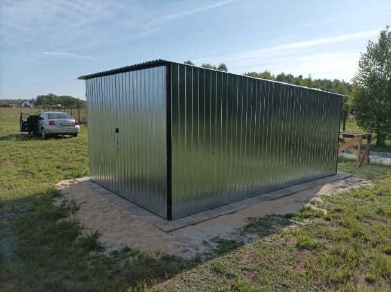Blaszany garaż z blachy trapezowej na betonowej wylewce, samochód z otwartymi drzwiami w tle, błękitne niebo.