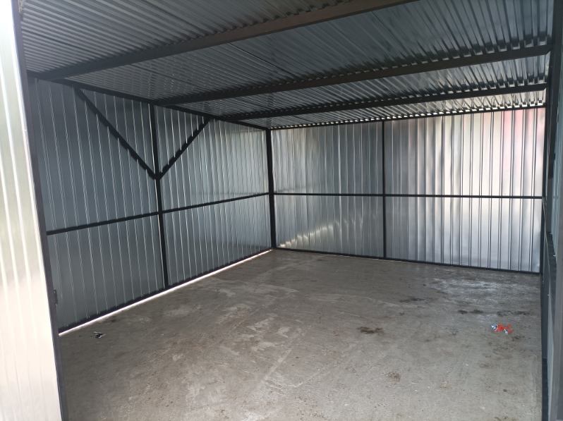 Wnętrze blaszanego garażu z widoczną konstrukcją stalową i pofałdowaną blachą. Surowy beton na podłodze. Ujęcie z wnętrza, podkreślające przestrzeń i solidność.