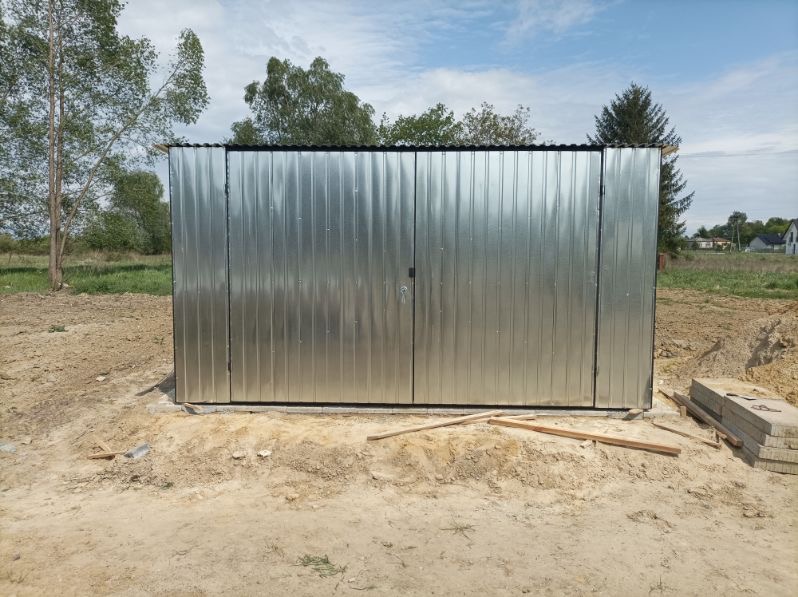 Garaż blaszany z dwuspadowym dachem w Poznań, z widocznymi śladami montażu i przygotowanym podłożem, na tle zieleni i błękitnego nieba.