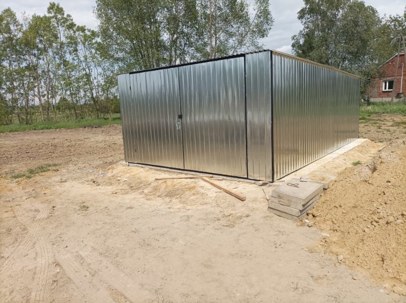 Metalowy garaż blaszany z dwuskrzydłowymi drzwiami, stojący na betonowej wylewce na piaszczystym podłożu, w tle drzewa i budynek mieszkalny.