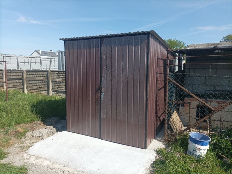 Brunatny garaż blaszany z dwuskrzydłowymi drzwiami, ustawiony na betonowej wylewce. W tle widoczne kontenery i ogrodzenie.