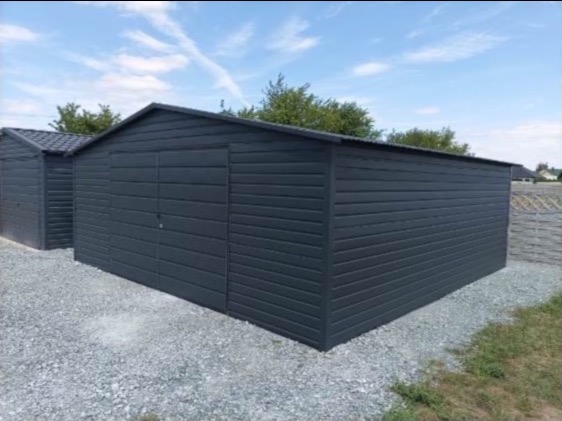 Dwa grafitowe garaże blaszane z poziomym panelowaniem, ustawione na żwirowym podłożu, w tle błękitne niebo i drzewa.