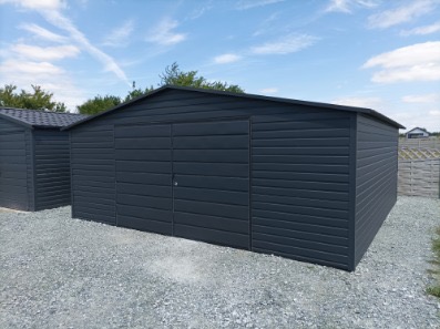 Grafitowy garaż blaszany z dwuskrzydłowymi drzwiami na żwirowym podłożu, widoczny drugi garaż w tle, błękitne niebo z chmurami.