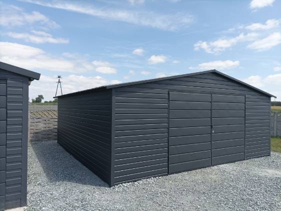 Grafitowy garaż blaszany z dwuskrzydłowymi drzwiami na żwirowym podłożu, widok z boku, na tle błękitnego nieba z delikatnymi chmurami.