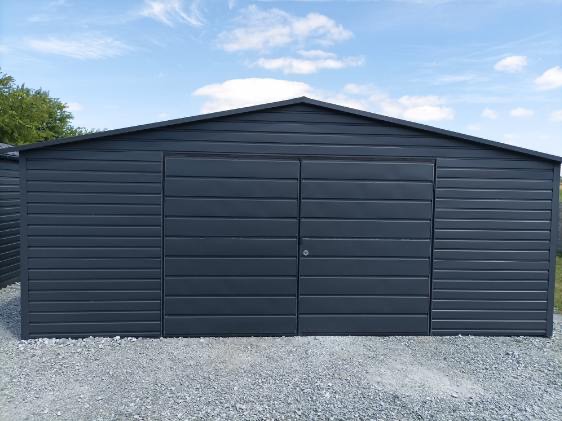 Grafitowy garaż blaszany z dwuskrzydłowymi drzwiami i pojedynczymi drzwiami bocznymi, ustawiony na żwirowym podłożu pod błękitnym niebem.
