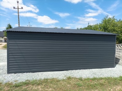 Grafitowy garaż blaszany z poziomym żłobkowaniem, na żwirowym podłożu, w słoneczny dzień. Widoczne elementy konstrukcyjne i trapezowa blacha na dachu.