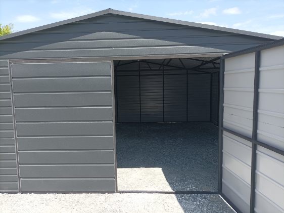 Otwarty, szary garaż blaszany z widocznym wnętrzem wypełnionym żwirem, prezentowany w jasnym świetle dziennym, z akcentem na konstrukcję i materiał.